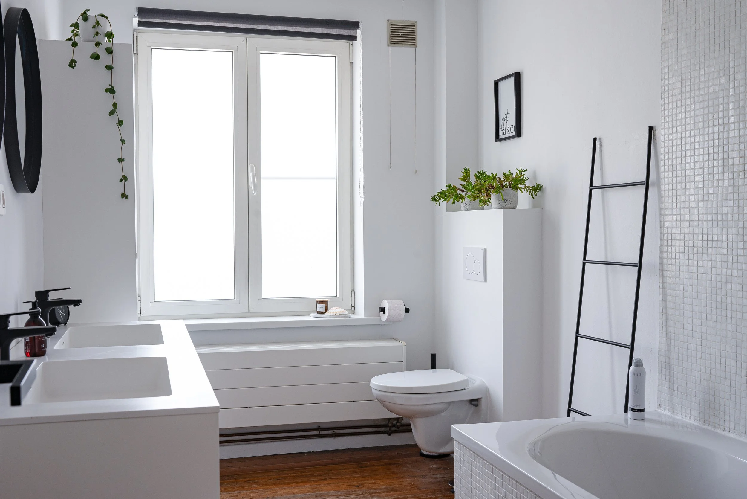 Modern bathroom with white walls, window, potted plants, black ladder, toilet, and bathtub.