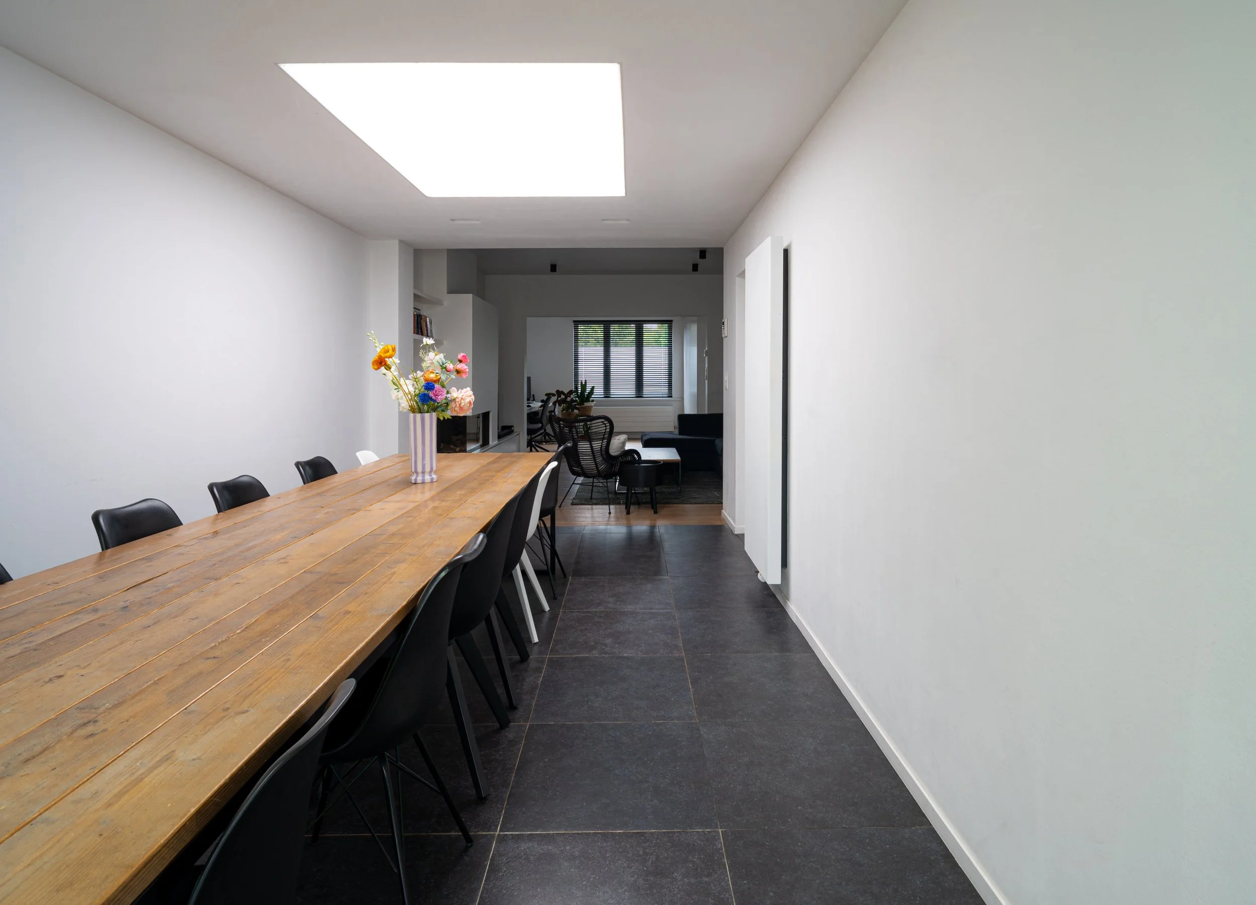 Modern dining room with white walls, a wooden table, black chairs, a vase with colorful flowers, and a living room area with a black sofa and window blinds in the background.