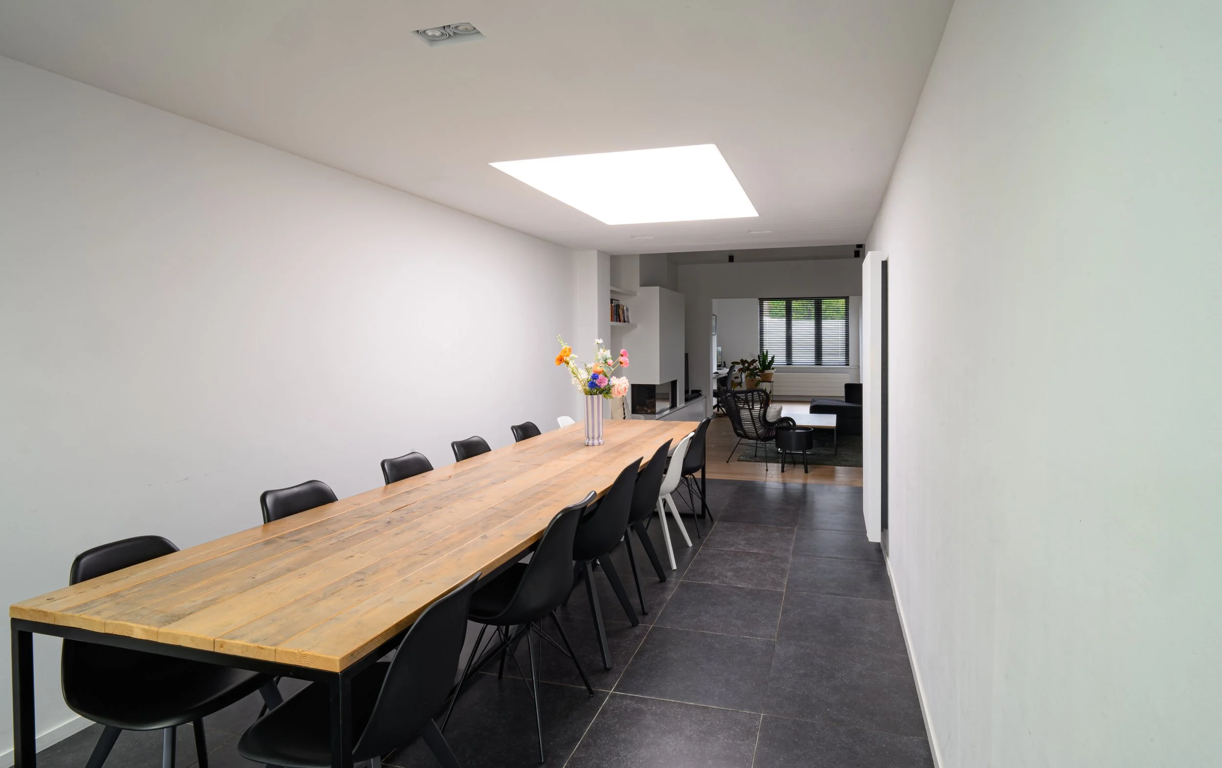 A modern dining area with a long wooden table surrounded by black chairs, a vase with colorful flowers, and a bright white ceiling light or skylight, with a living space visible in the background.