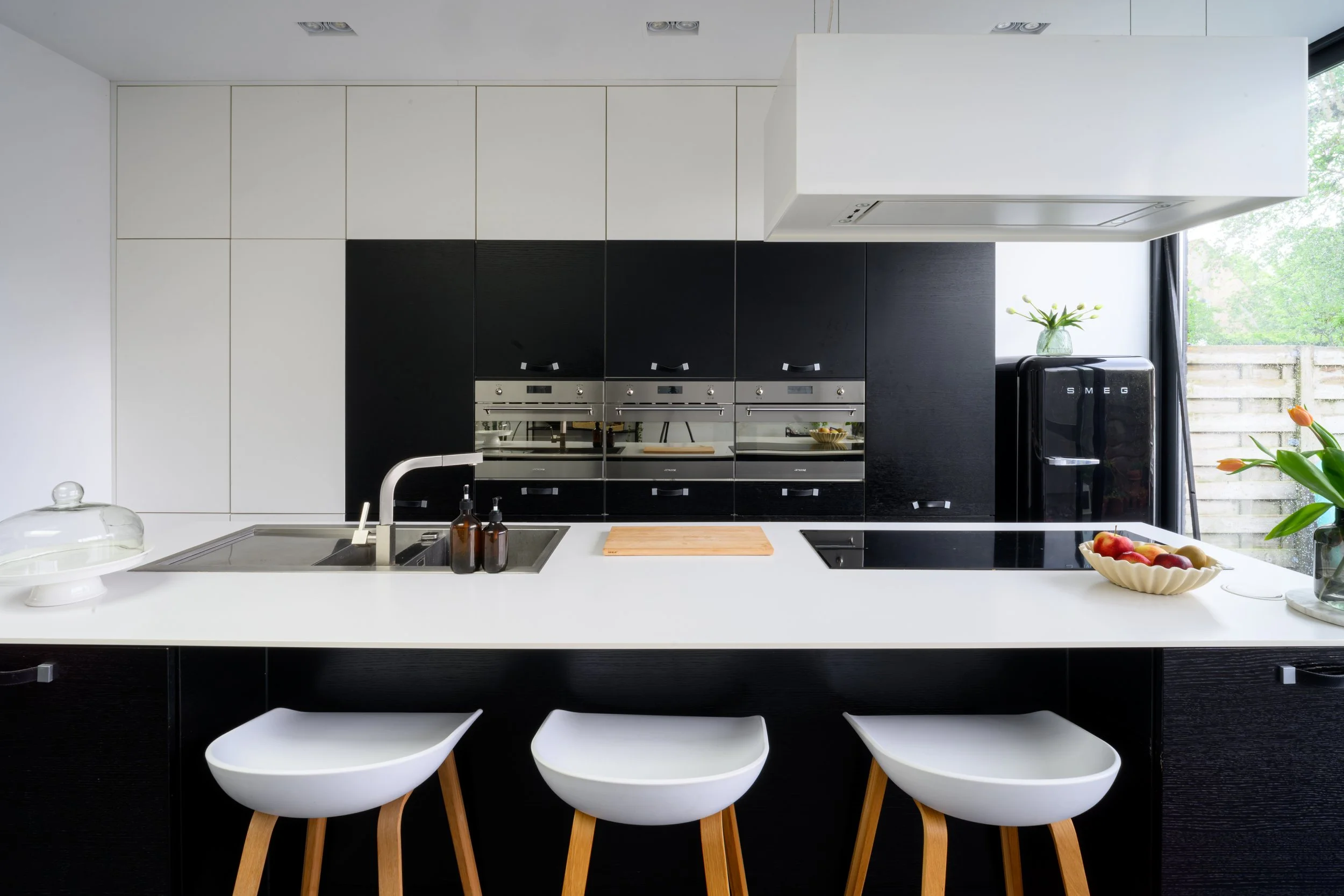 Modern kitchen with black and white cabinets, a white countertop, and white chairs. A window with a view of green trees, a black Smeg fridge, and decorative items like a vase with flowers and bowls.