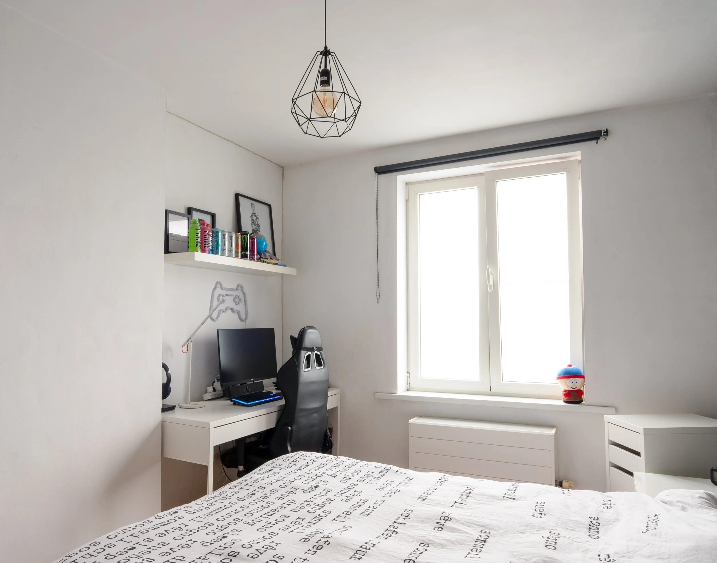 A bedroom workspace with a white desk, a monitor, a black ergonomic chair, and a black gaming chair. The desk has a strip of colorful cans and framed artwork. There is a window with a black curtain rod and a plush toy on the windowsill. A geometric b