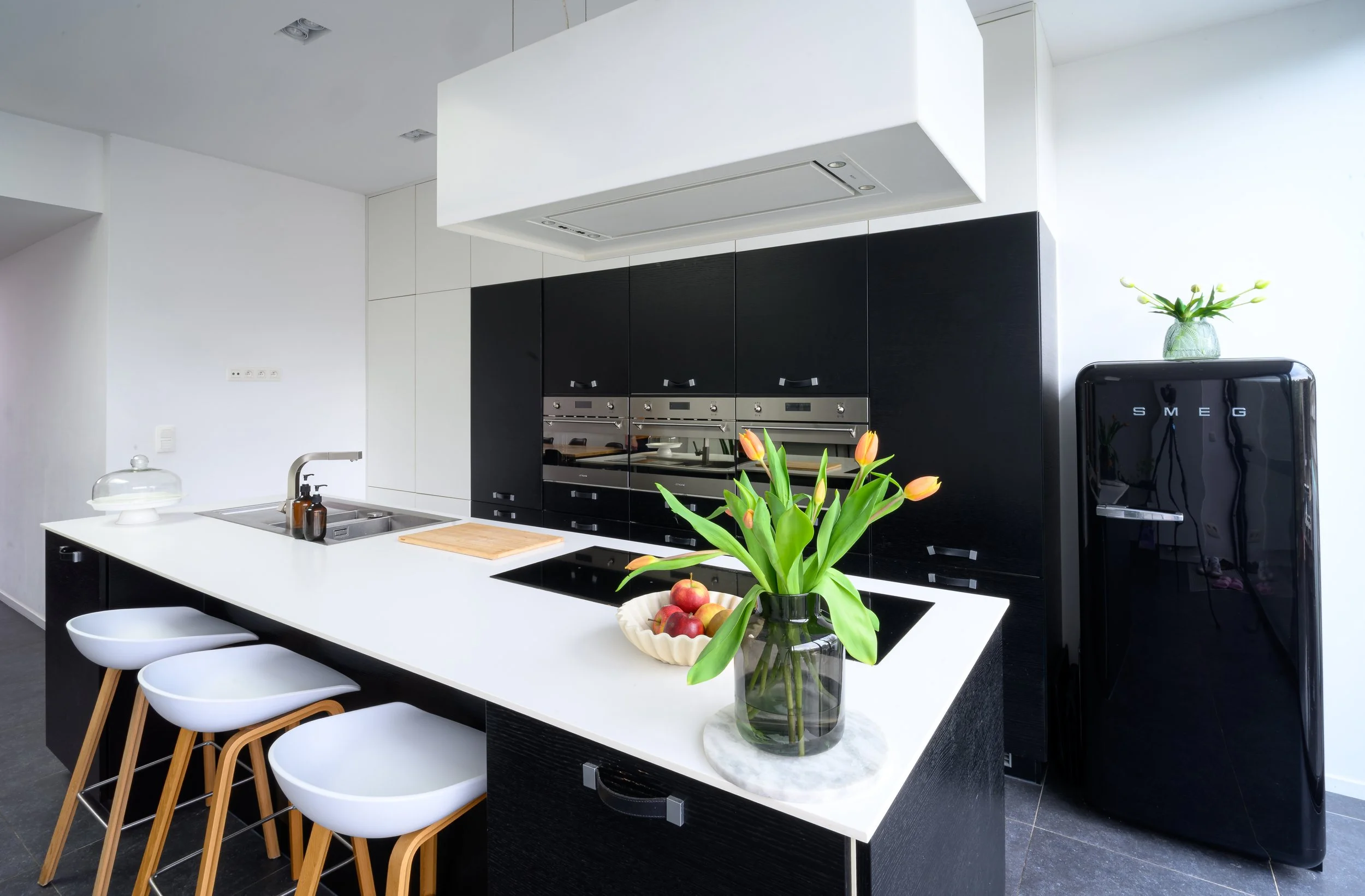 Modern kitchen with black and white cabinetry, a white island with seating, a vase with tulips and apples, and a black SMEG refrigerator.