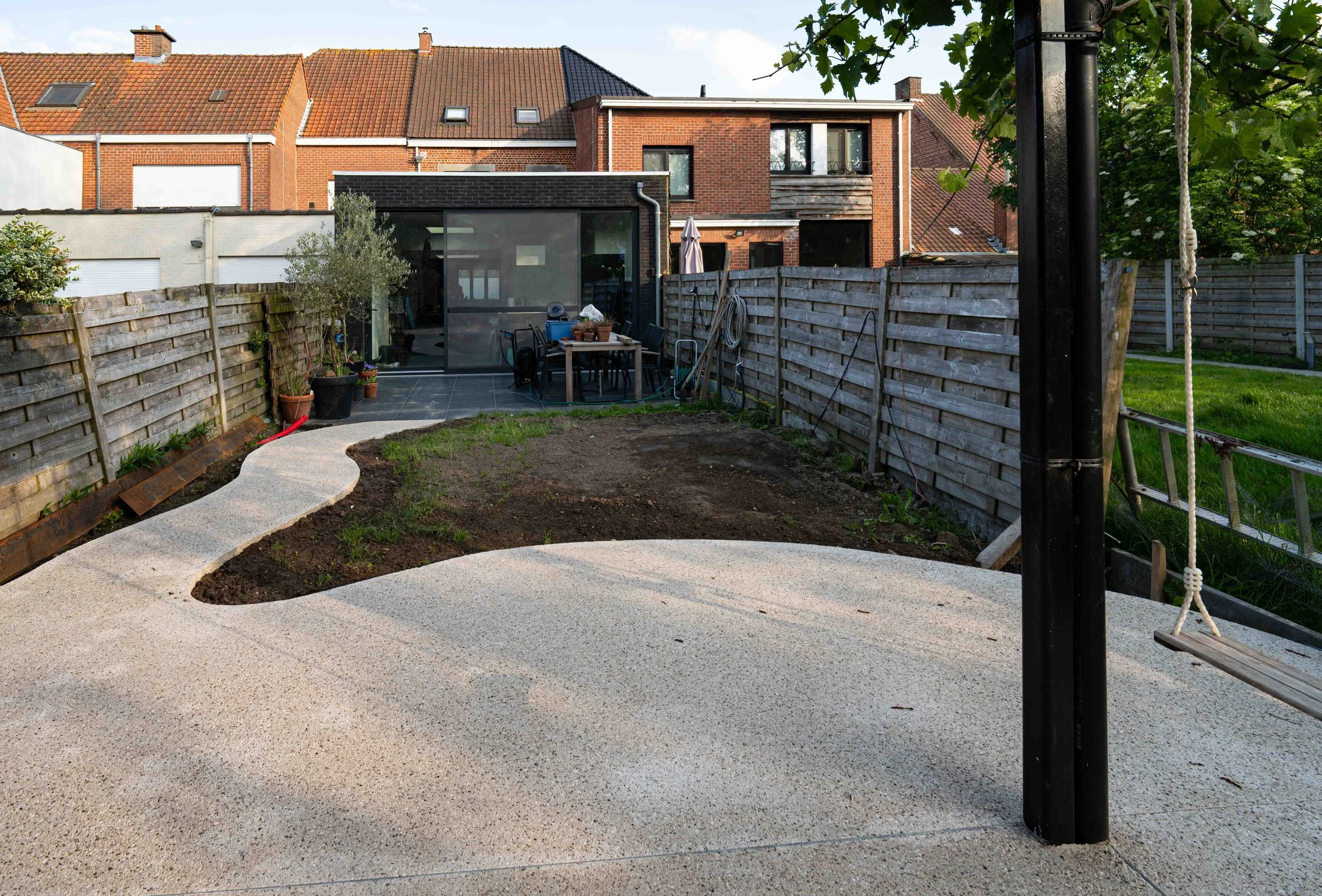 A backyard with a newly poured concrete patio, a wooden fence, potted plants, and a modern two-story brick house with a small balcony.