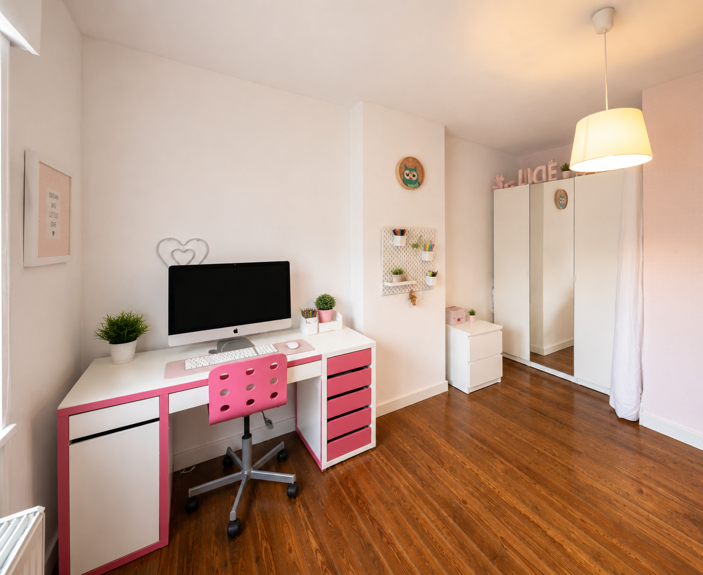 A modern bedroom with a white and pink desk, computer, pink chair, and wooden flooring. There is a wardrobe and decor with owl motifs, a wall-mounted shelf with plants, and a ceiling lamp.