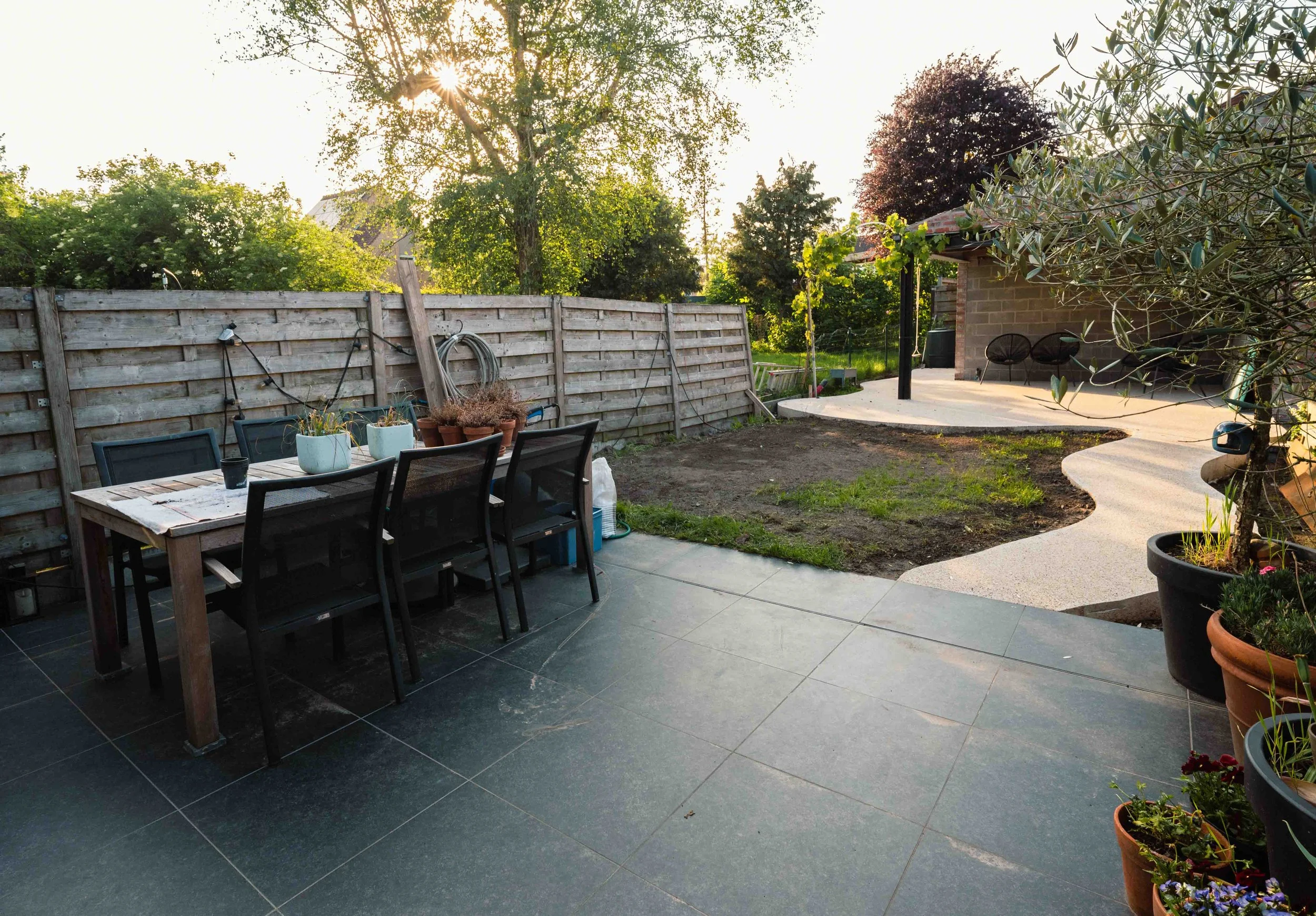 A backyard patio with a black outdoor dining table and six matching chairs, several potted plants, a weathered wooden fence, and a concrete pathway leading to a grassy yard with trees and bushes, illuminated by sunlight.