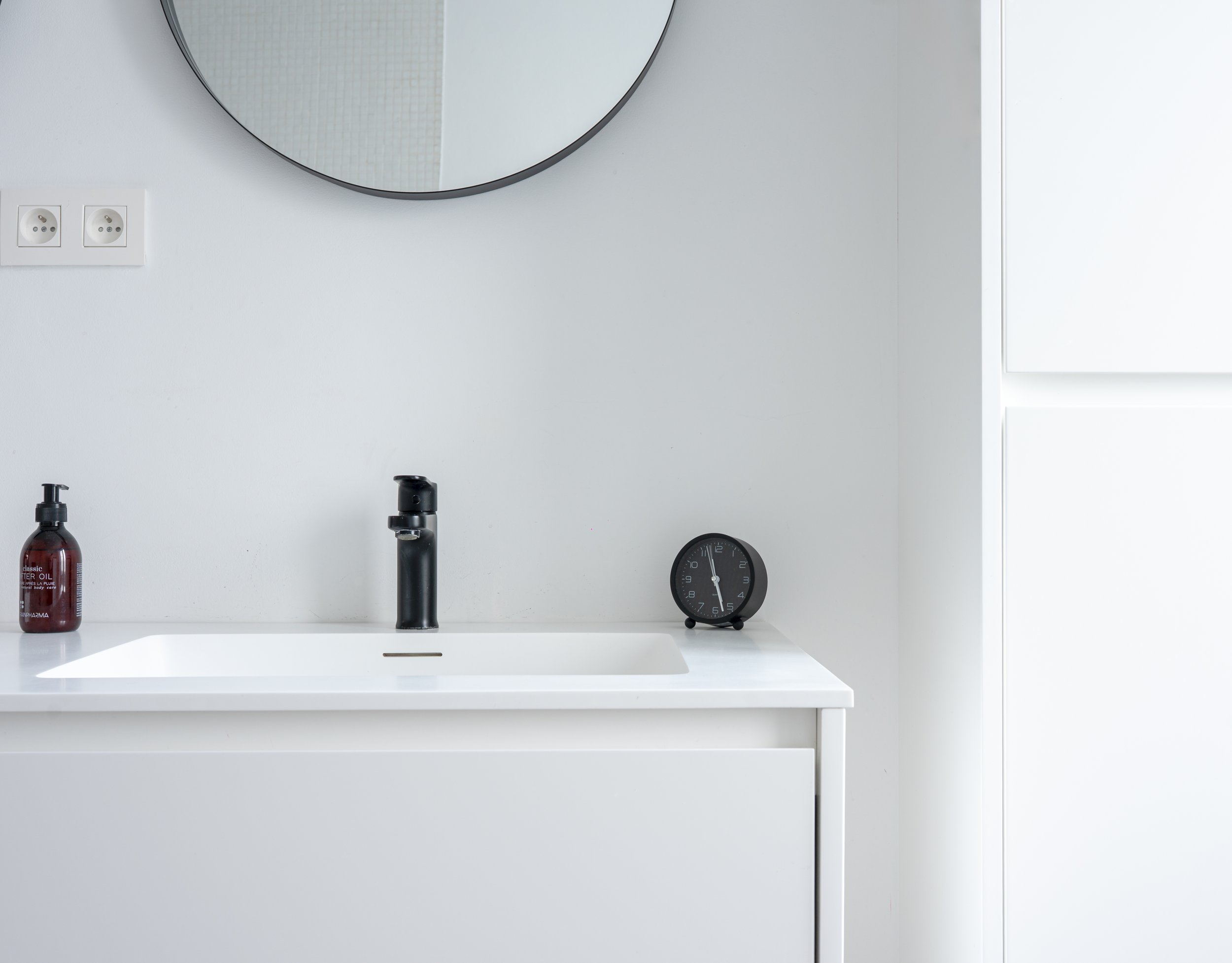 Minimalist bathroom sink area with a round mirror, two electrical outlets, a black soap dispenser, and a black faucet, with a white countertop and light gray wall.