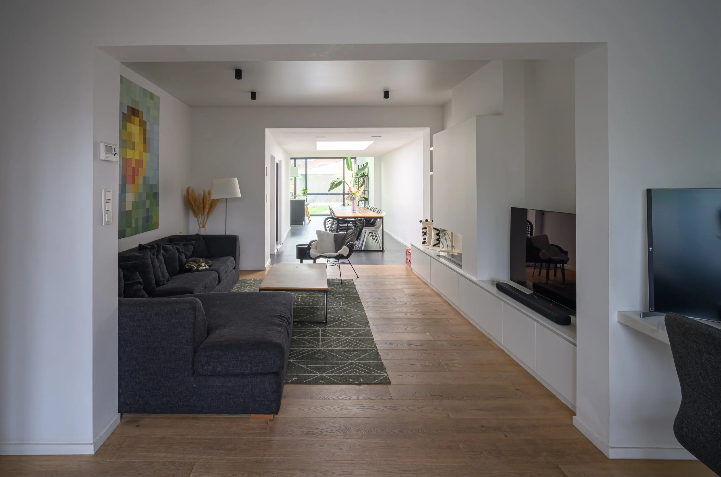 Living room with black sofa, wooden floor, wall art, and dining area in background with large windows and plants.