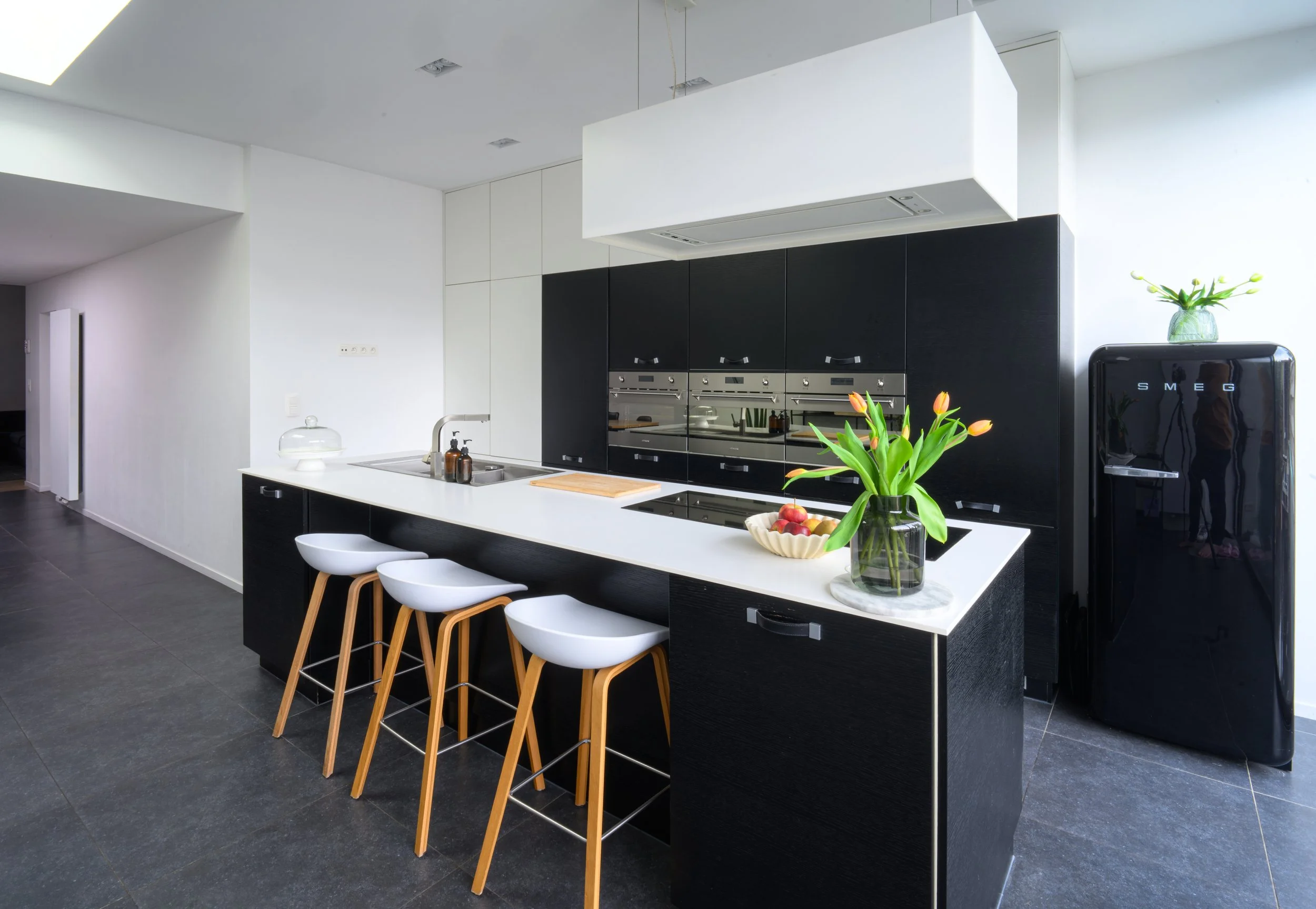 Modern kitchen with white cabinets, black accents, a white countertop island with four white stools, a bowl of fruit, a vase with tulips, and a black Smeg refrigerator.