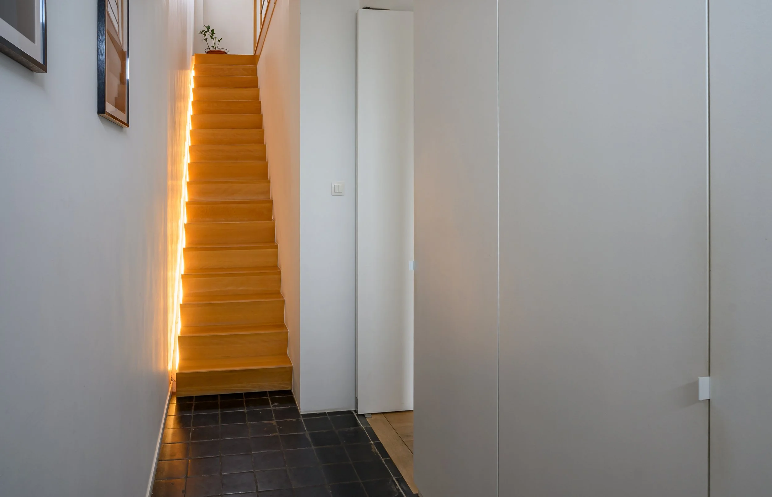 Interior view of a home, showing a wooden staircase with warm lighting along the side, next to white walls and a small corner with a potted plant at the top of the stairs. There are a few framed pictures on the wall to the left, and the foreground ha