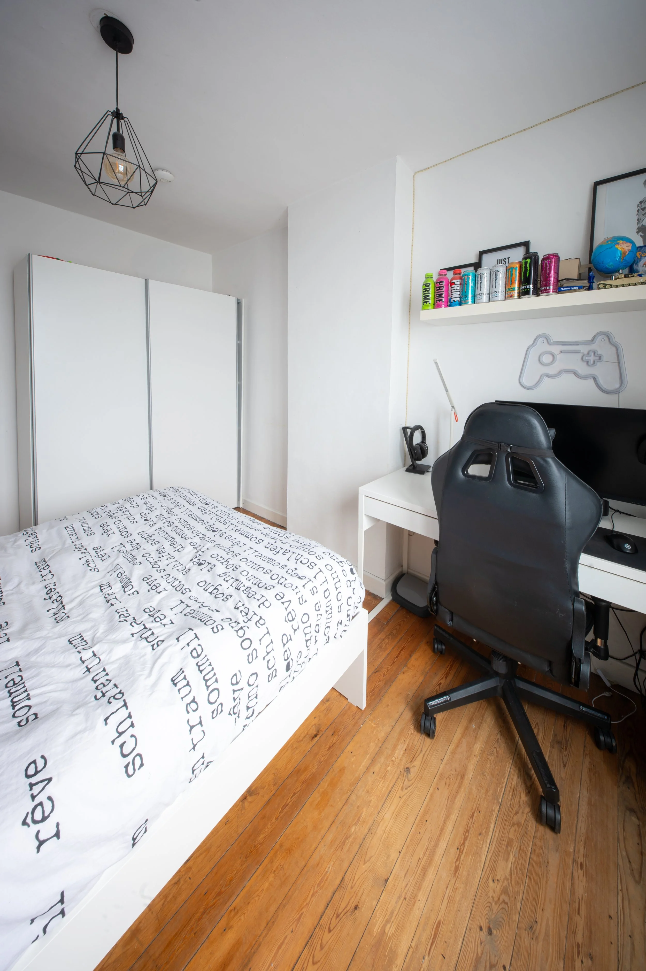 A bedroom with a white bed covered in a white blanket with black words, a white wardrobe, a desk with a black gaming chair, a shelf with cans, a globe, and picture frames, and a modern ceiling light fixture.