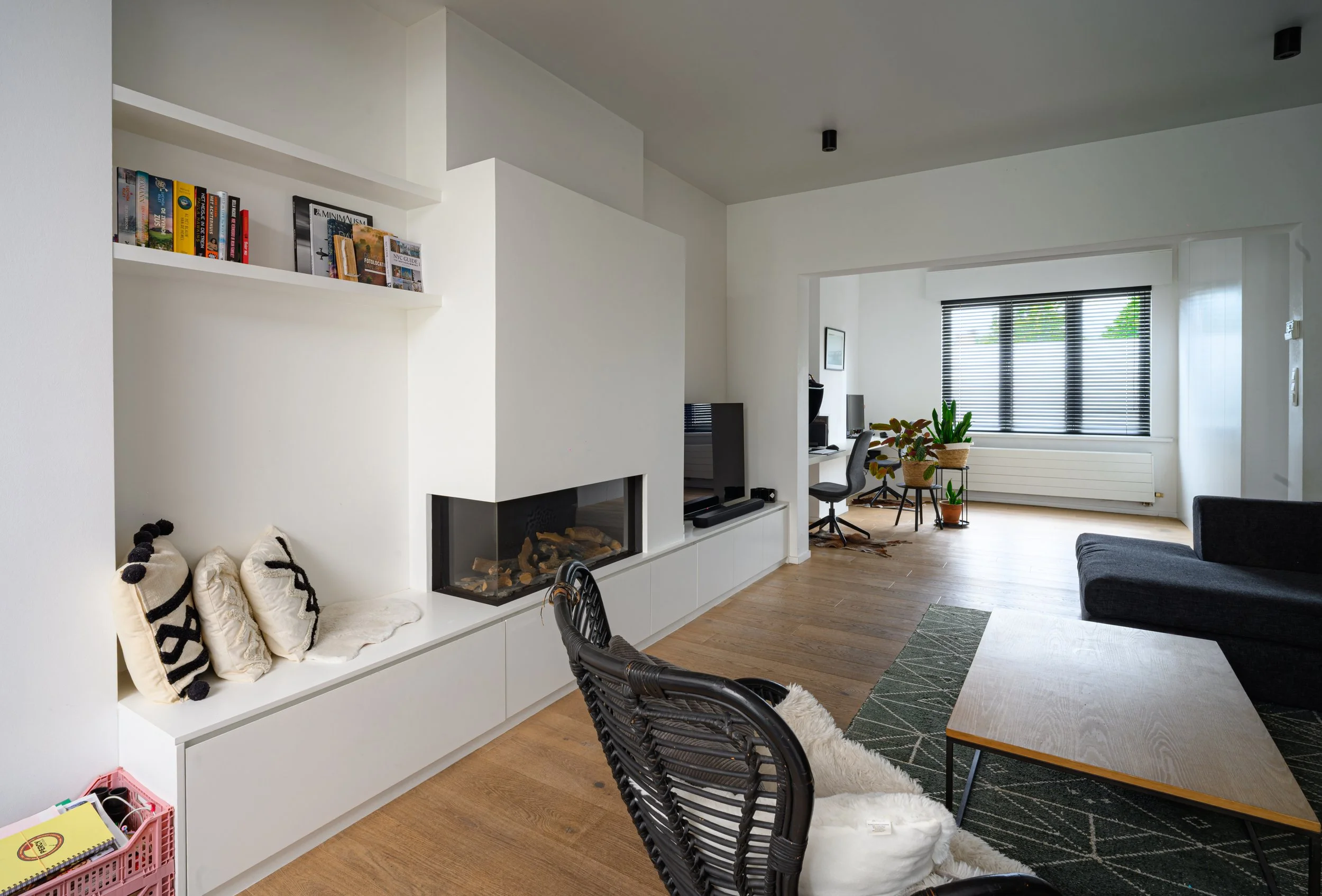 A modern living room with white walls, a built-in white fireplace with a glass enclosure, and a white shelf above it holding magazines and books. There are black and white throw pillows on the white built-in seating area. The room has wooden flooring
