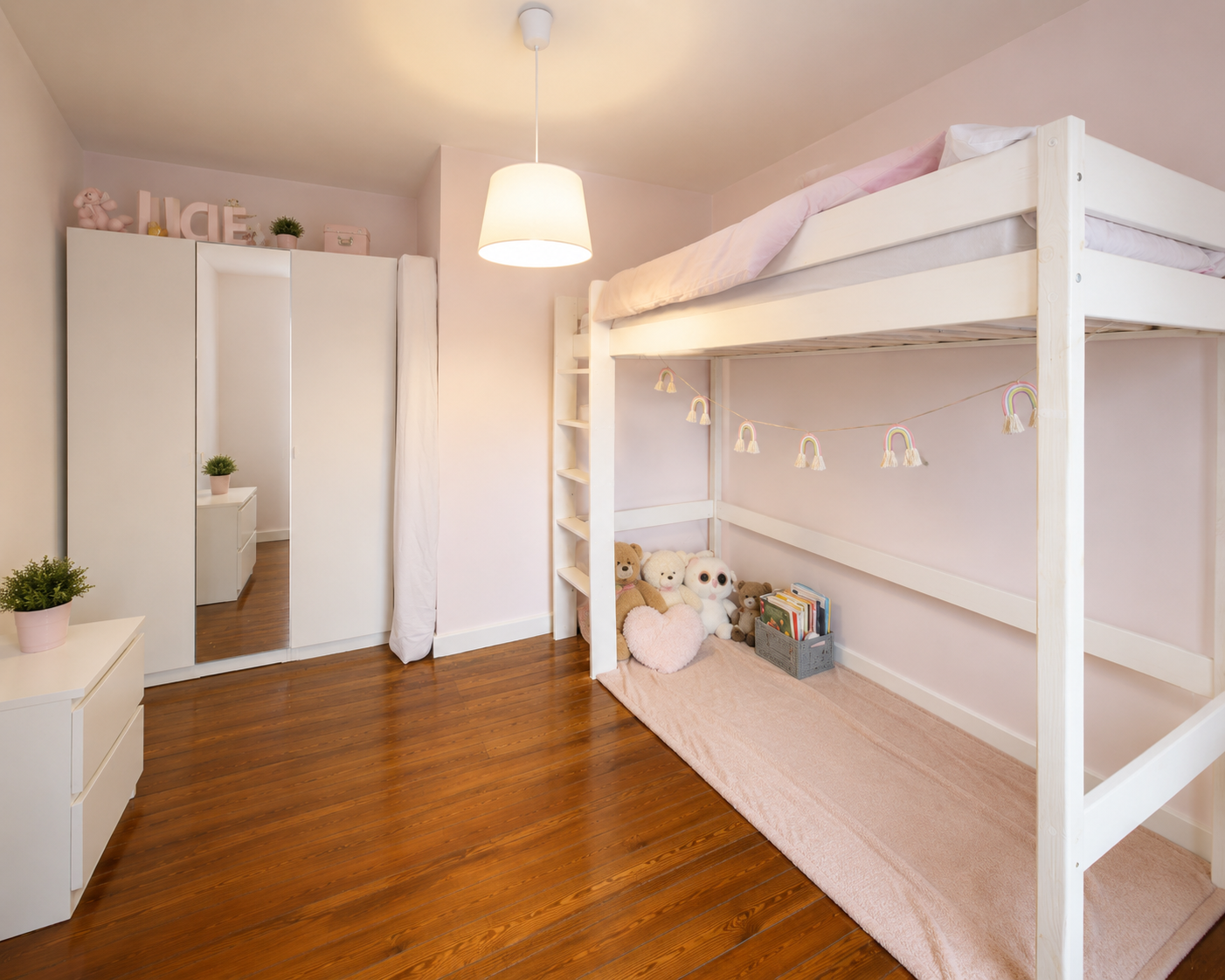 A children's bedroom with a white bunk bed and pink accents, including plush toys, a heart-shaped pillow, and a string of rainbow and rainbow-shaped decorations. There is a white wardrobe with a mirror and a white dresser with plants. The room has ha