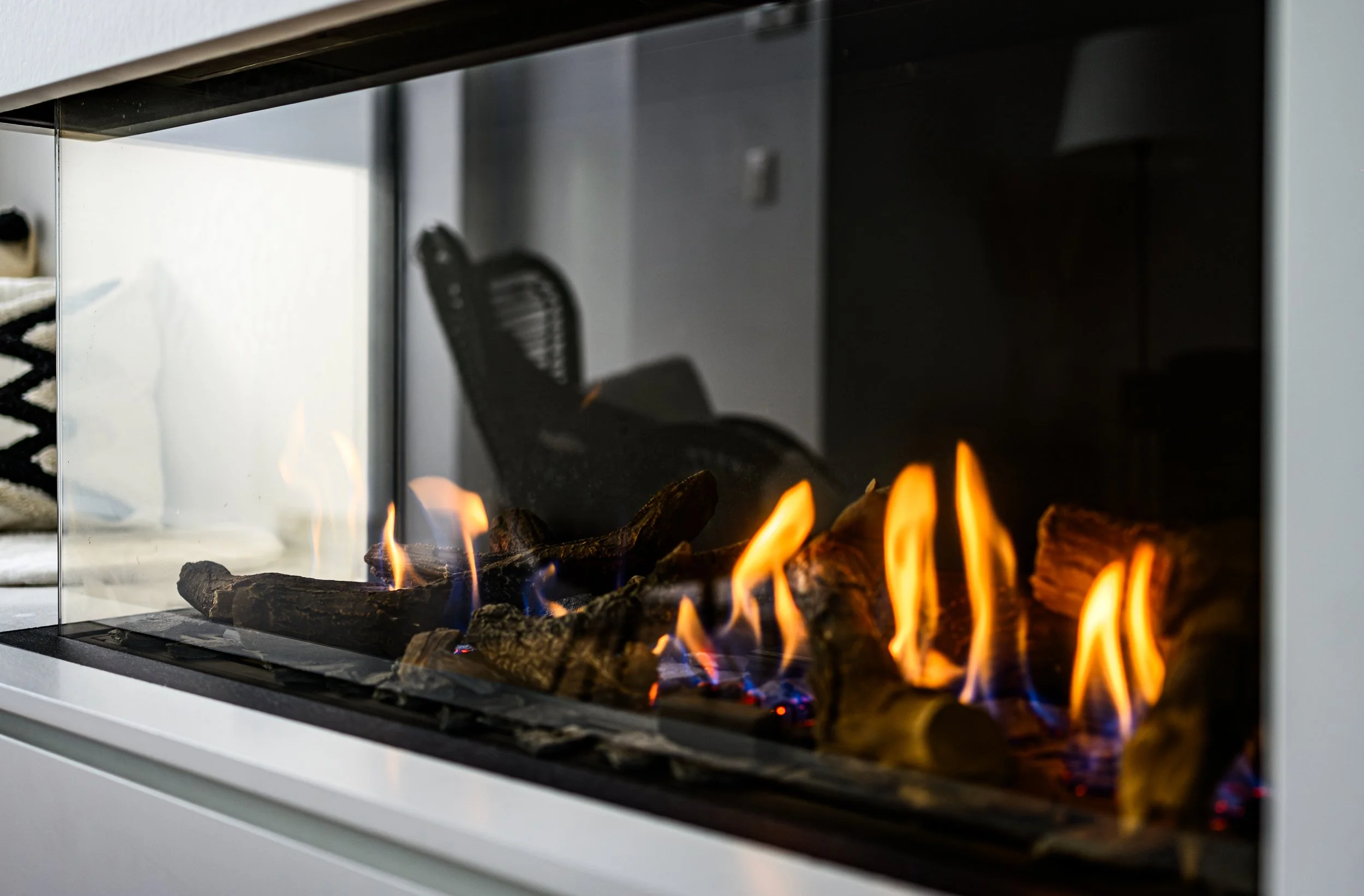 A modern fireplace with a glass front, showing flames and burning logs inside.