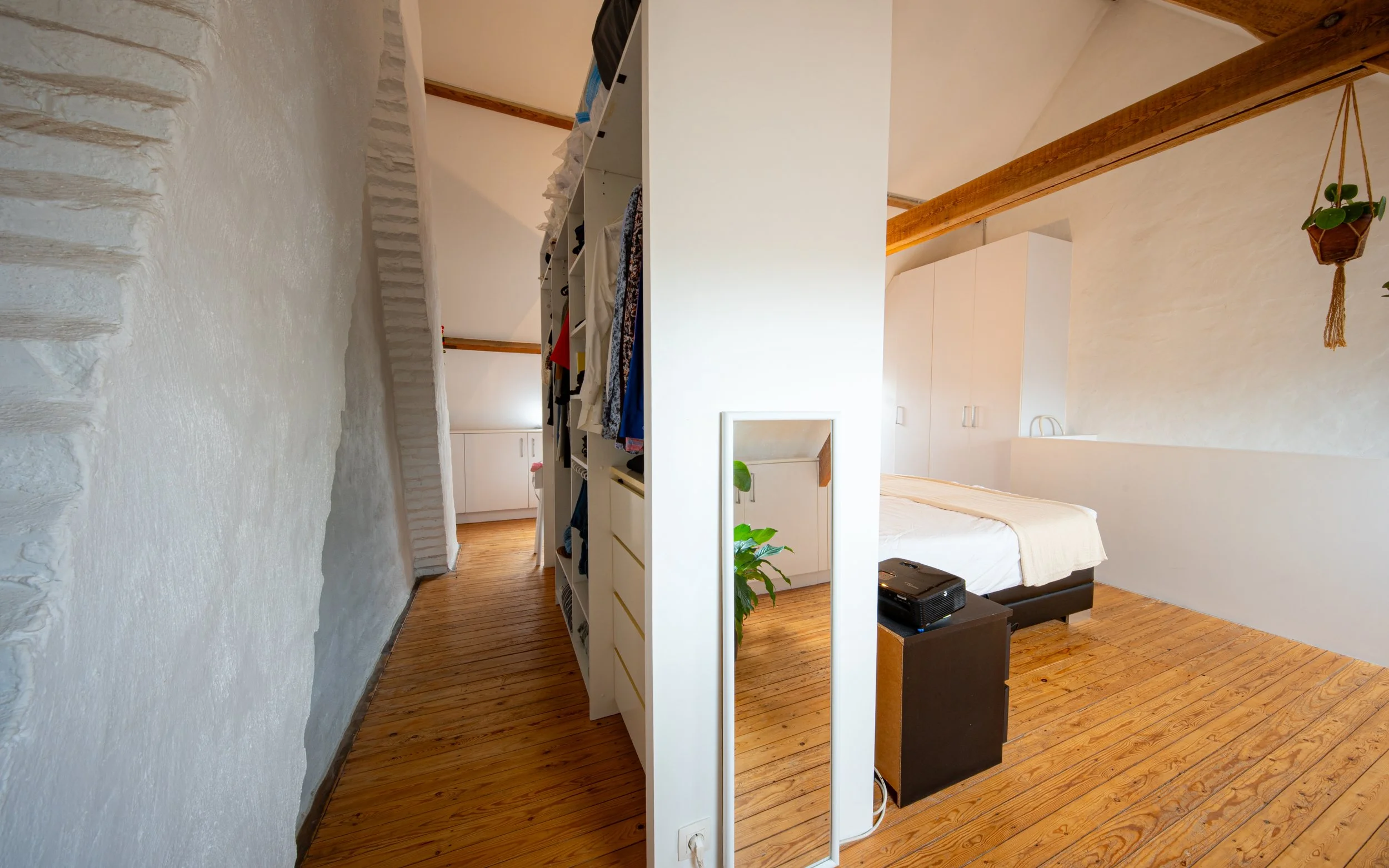 A cozy bedroom with white walls and wooden ceiling beams. There is a white wardrobe and an open closet with hanging clothes. A bed with a white bedspread and a black side table with a projector on it are also visible. A potted plant is placed on the 