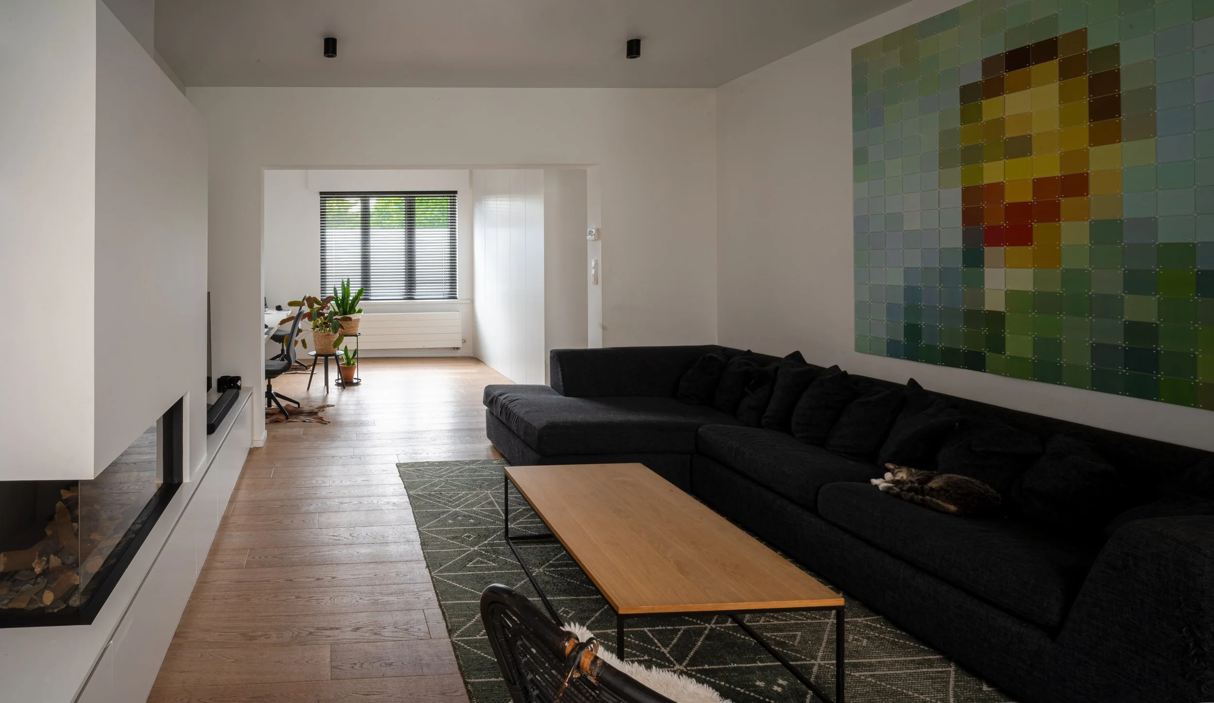Living room with black sofa, wooden coffee table, wall art made of colored tiles, and a fireplace, with a dining area with plants and window in the background.