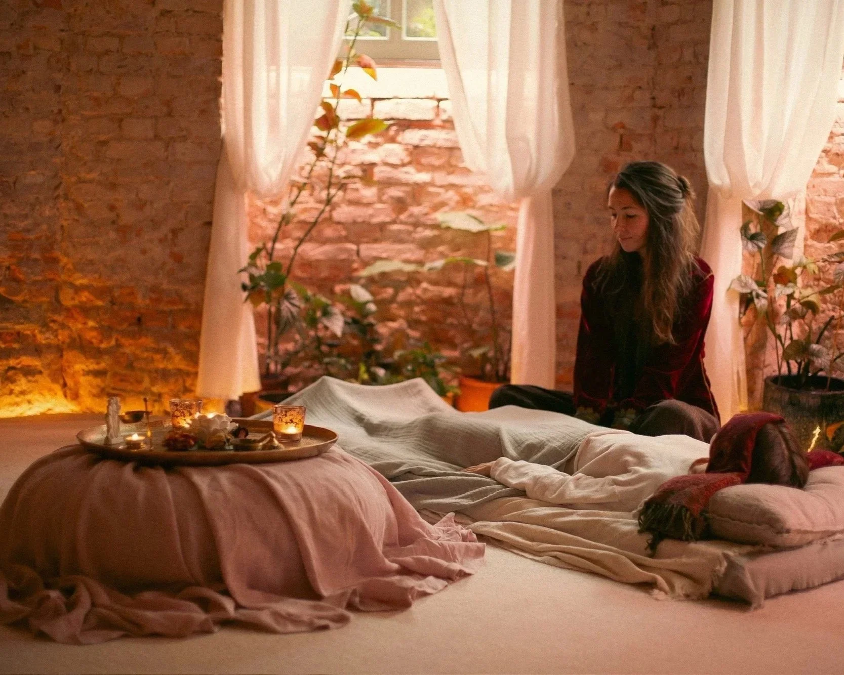 A woman with long dark hair sitting cross-legged indoors on a cozy bed with beige and pink blankets. There are candles and a tray of snacks on the bed. Sunlight filters through a window with white curtains, illuminating the brick wall and potted plants behind her.