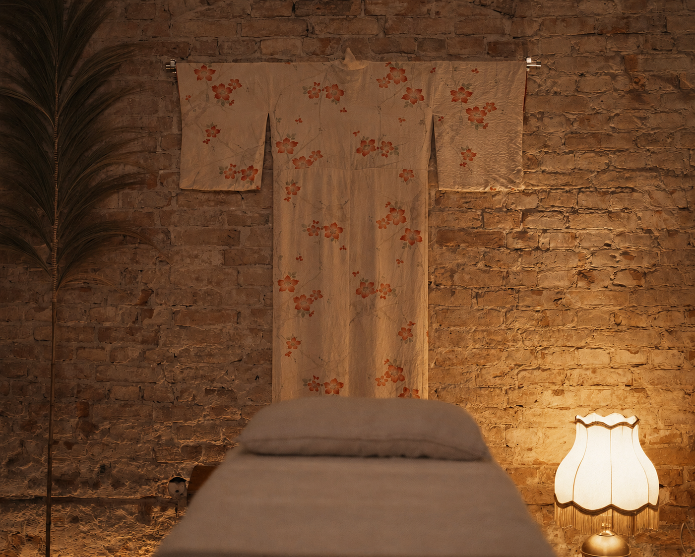 Interior of a cozy room with an exposed brick wall, a fabric wall hanging with floral patterns, a tall indoor plant, and a vintage table lamp providing warm lighting.