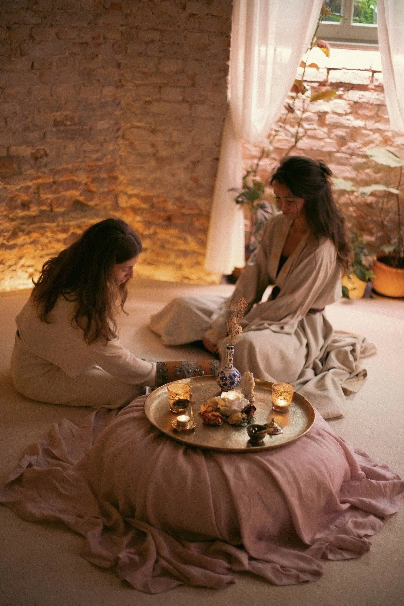 Two women dressed in beige robes sitting on the floor around a low, pink, flower-shaped cushion with a gold tray of snacks, candles, and a vase with flowers on top. The setting appears cozy with a brick wall, a window with curtains, and plants in the background.