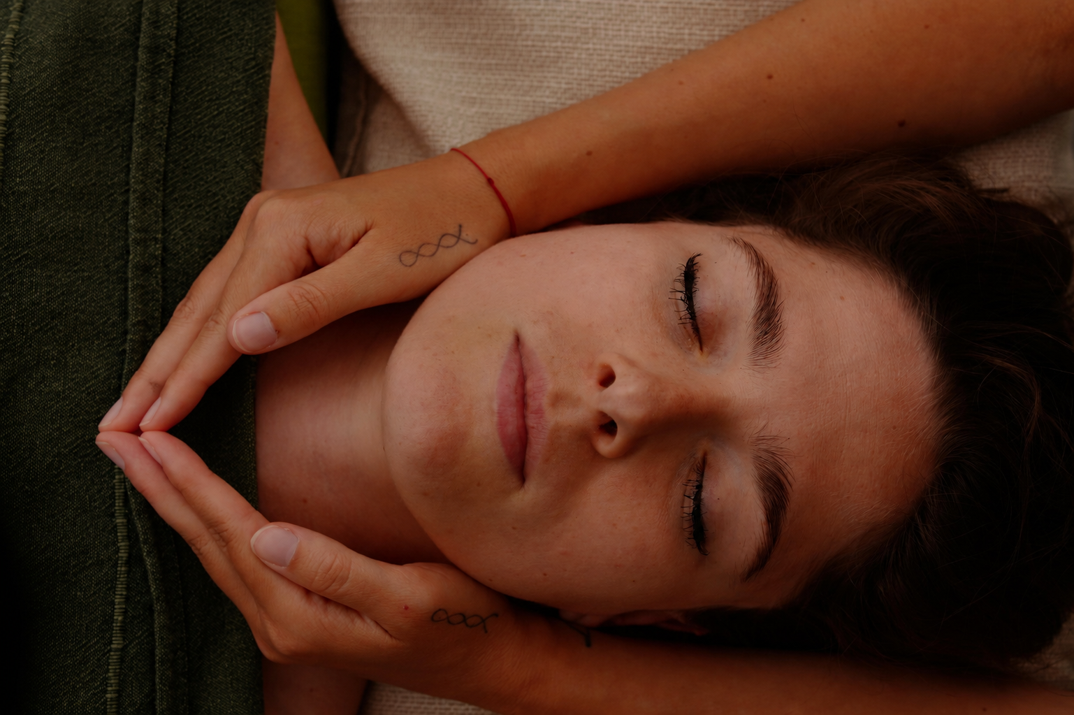 A woman with closed eyes is lying down with her hands gently framing her face. She has freckles, dark eyebrows, and long eyelashes, and appears relaxed.