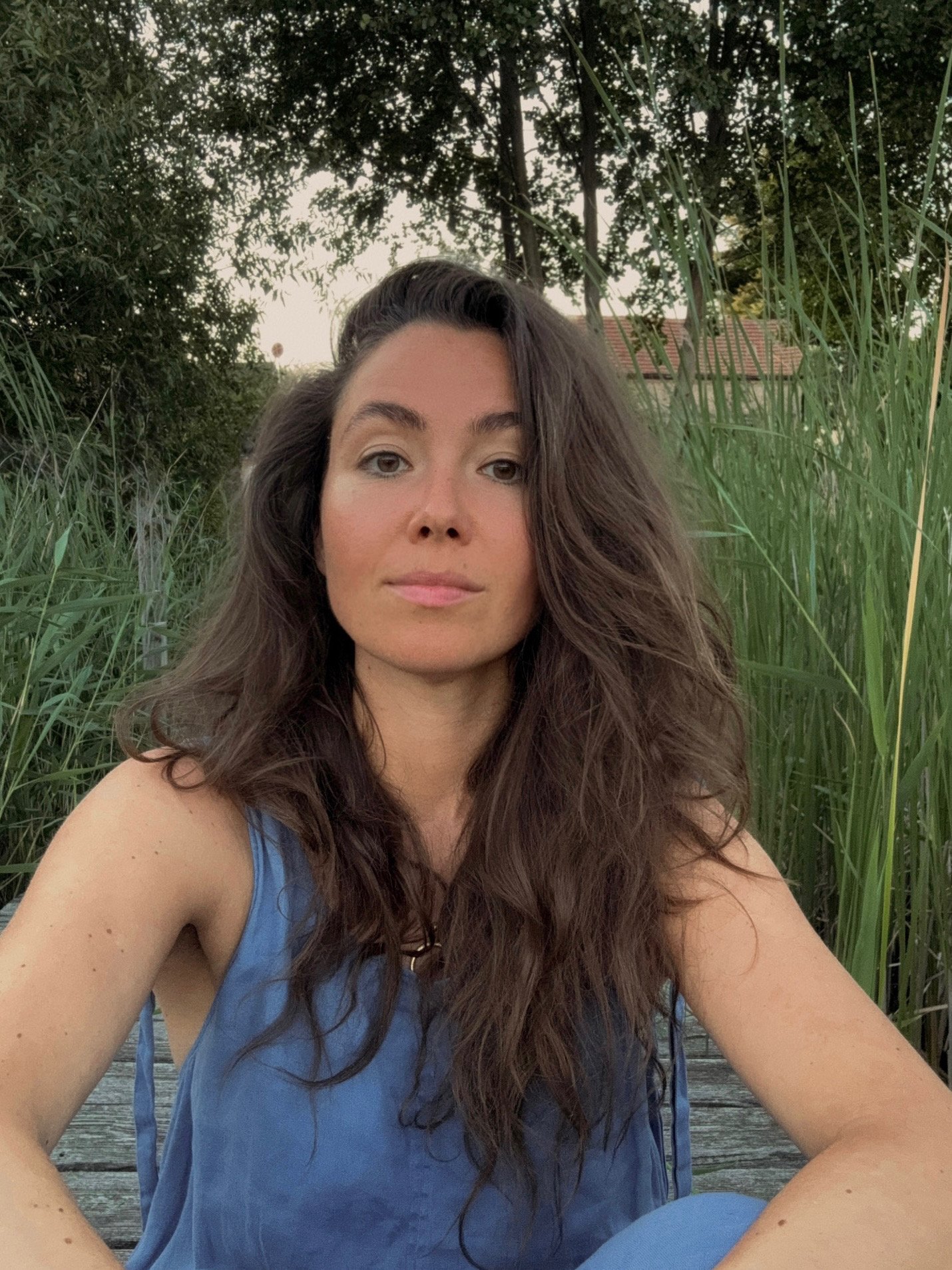 A woman with long brown hair sitting in a natural outdoor setting with green plants and trees behind her, wearing a blue sleeveless top, looking at the camera.
