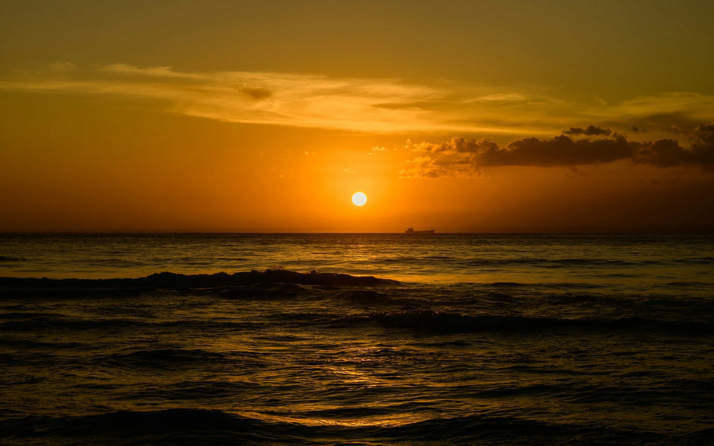Sunset over the ocean with a boat visible near the horizon, partly cloudy sky with golden light.