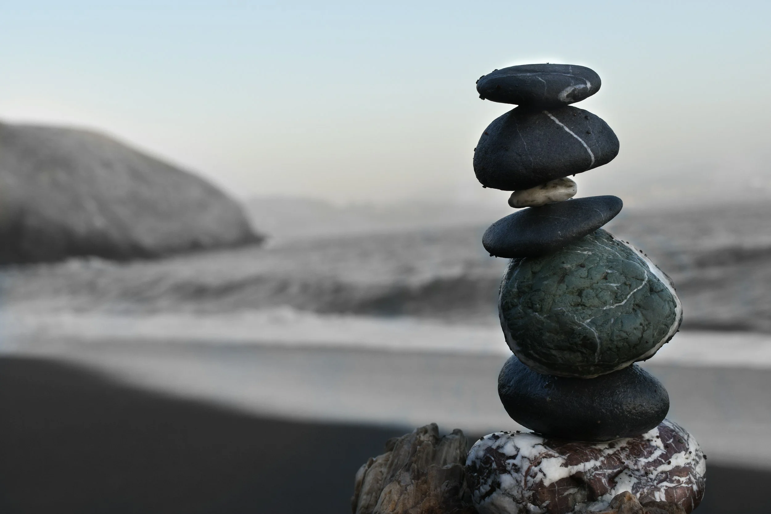 Stacked smooth black and gray stones on a beach with blurred rocks and ocean in the background.