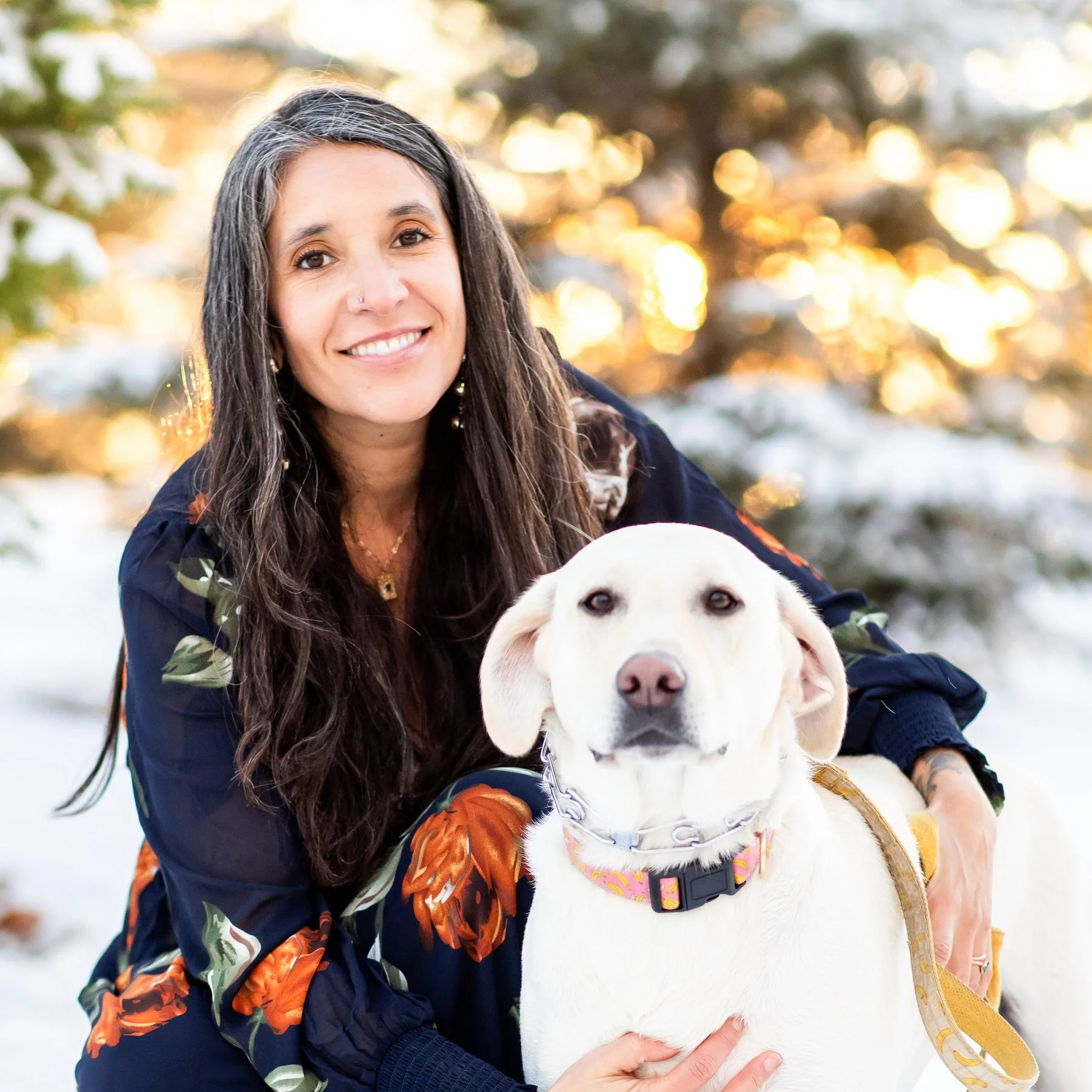 A woman with long dark hair smiling and holding a white Labrador retriever puppy outdoors during winter, with snow on the ground and trees in the background, illuminated by warm sunlight.