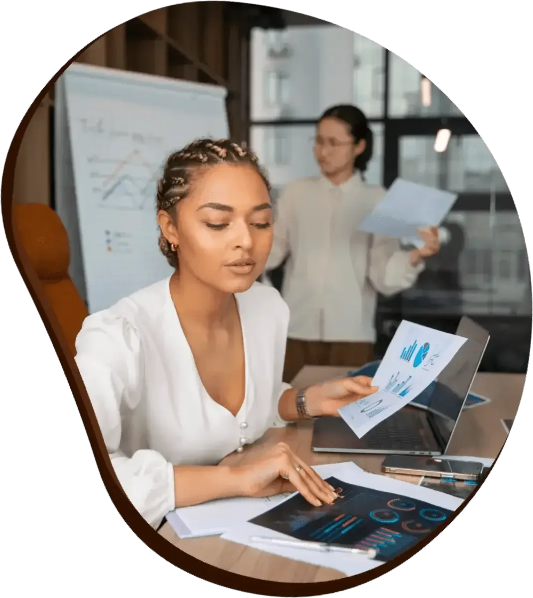 Two women working on business documents and charts in a modern office with large windows, one sitting at a desk and the other standing with papers.