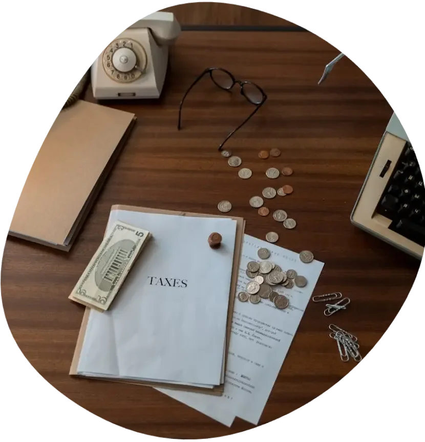 An overhead view of a wooden desk with bookkeeping items including a calculator, glasses, coins, a tax document, a paperclip, and a five-dollar bill.