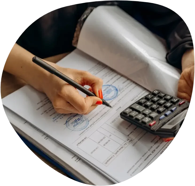 Person signing documents with a pen, holding a calculator, and reviewing papers on a wooden desk.