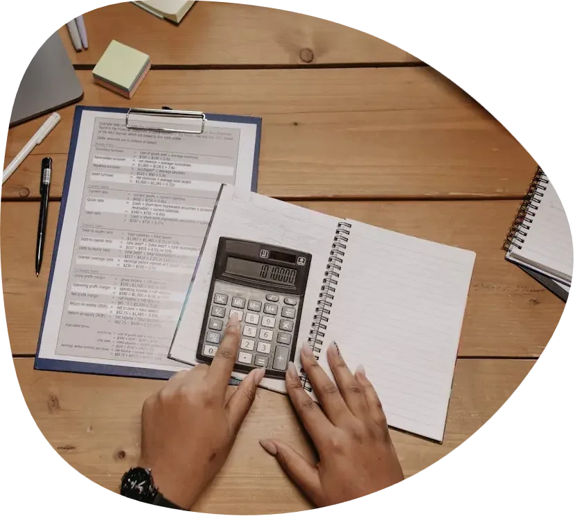 Person using a calculator on a wooden desk with notebooks, a clipboard with documents, pens, and sticky notes.