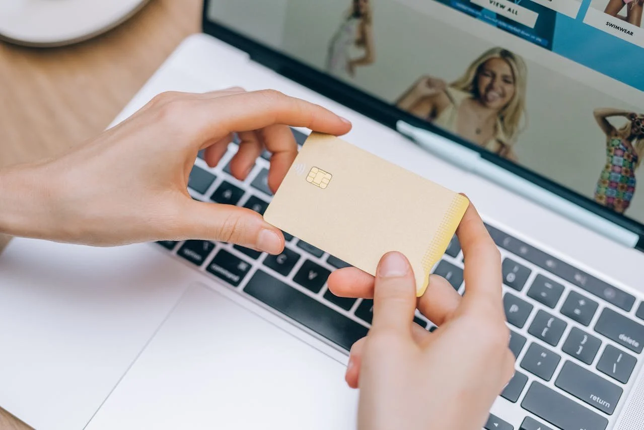 Person holding a credit card in front of a laptop showing a fashion website.