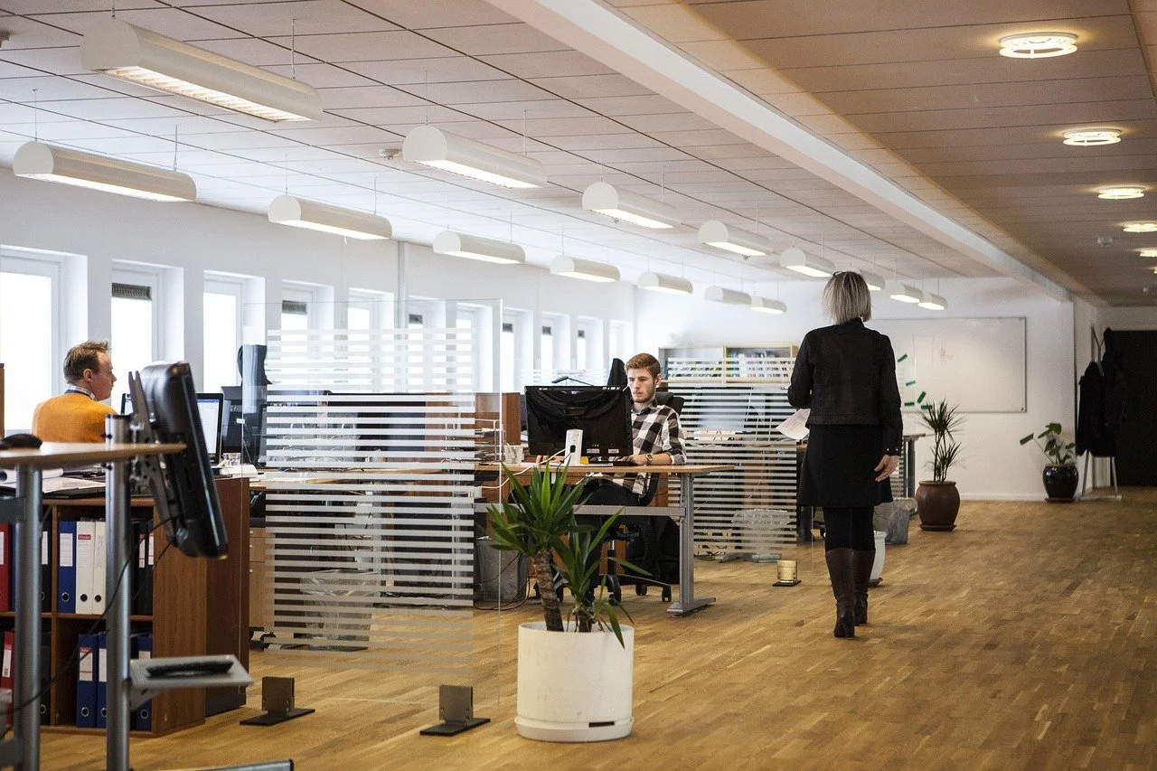 An office with multiple workstations separated by glass partitions, a woman walking toward a seated man at a desk, and various potted plants and office supplies, under ceiling lights and windows.