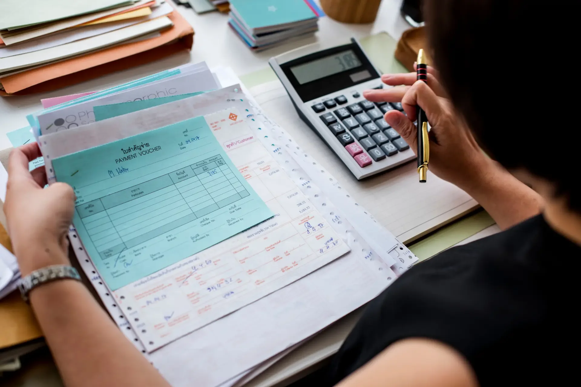 Person working at a desk with a calculator, notebooks, and a payment voucher in hand, surrounded by stacks of paper and files.