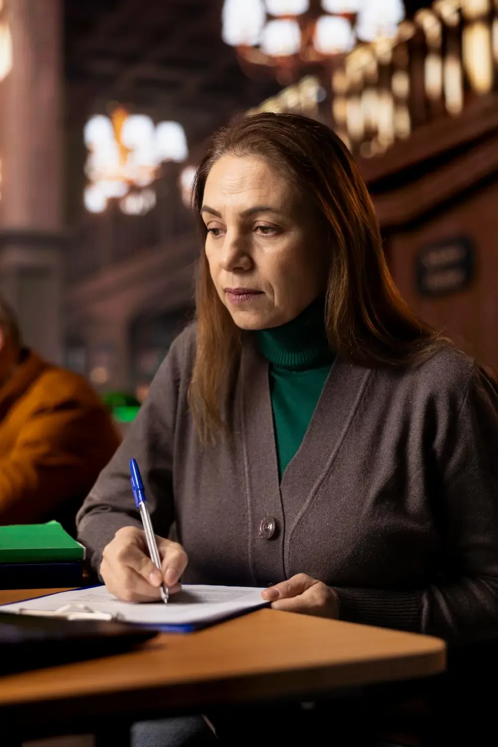 A woman in a brown jacket writing with a blue pen on a notepad at a wooden table, in a dimly lit environment with wooden interior and blurred background.