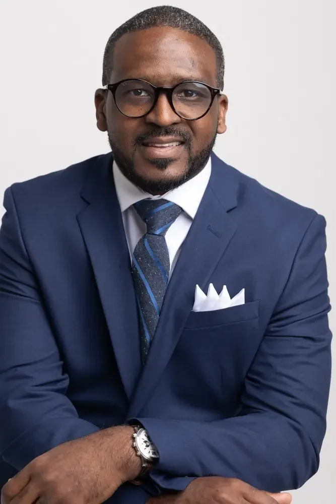 A professional man with glasses, wearing a navy blue suit, white dress shirt, and a striped tie, with a white pocket square, posing against a plain white background.
