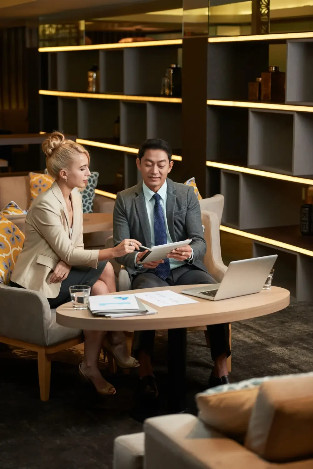 Two business professionals, a man and a woman, having a discussion and reviewing documents in a modern, well-lit lounge area with a laptop, papers, and a glass of water on the table.