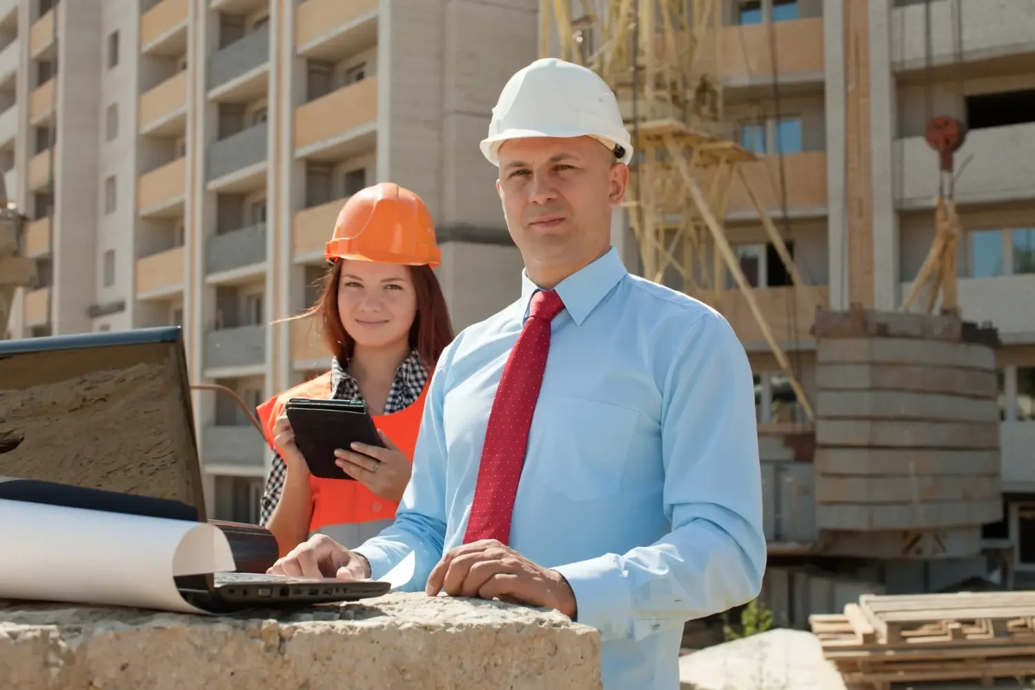 Two construction professionals, a man in a white hard hat and a woman in an orange hard hat, are on a construction site with a building under construction in the background. The man is working on a laptop, and the woman is holding a tablet, both dressed in professional attire suitable for construction supervision.