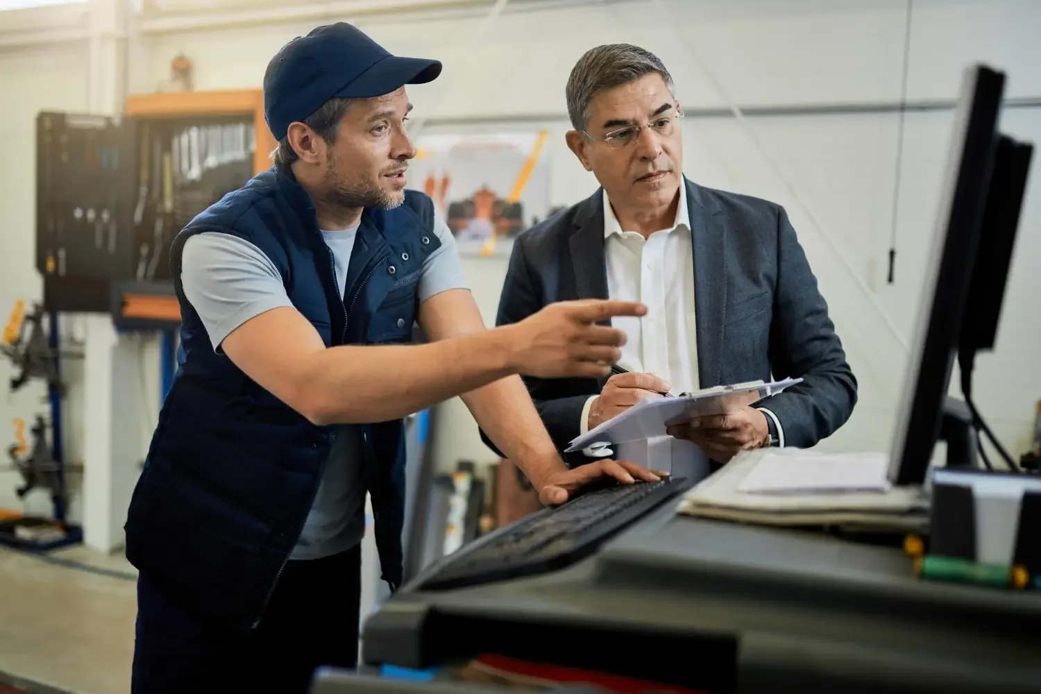 Two men are working together in an industrial or workshop setting, looking at a computer screen and discussing something. One man is pointing at the monitor and holding a clipboard, while the other is wearing a cap and casual work attire. There are tools and equipment in the background.