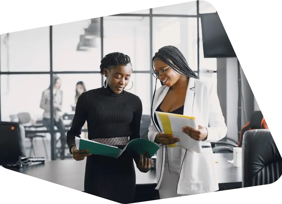 Two women are discussing documents in a modern office with large windows and a conference table in the background.