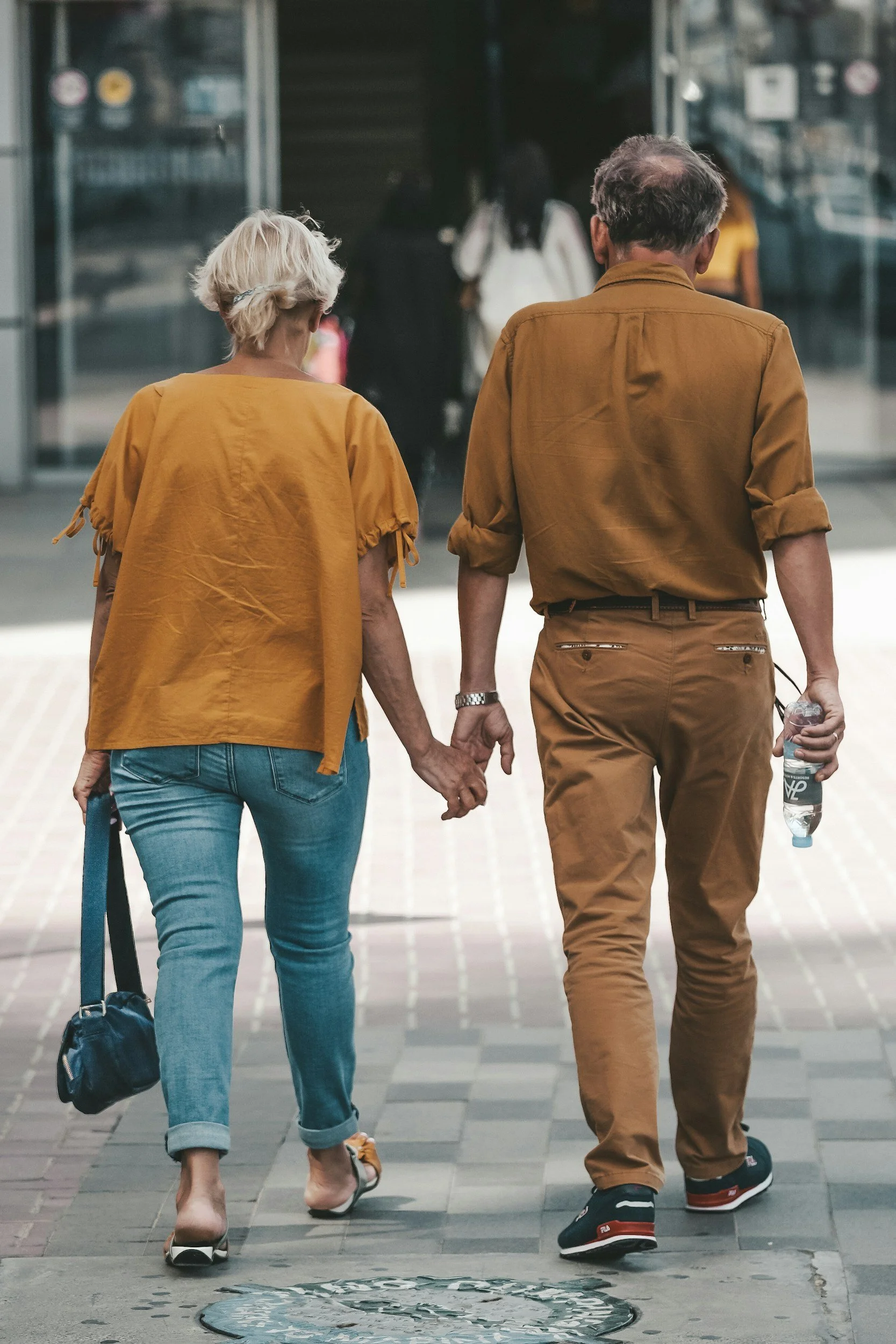 A middle-aged couple walking hand-in-hand on a city sidewalk, with the woman wearing ripped blue jeans and a yellow blouse, and the man in brown pants and a shirt, holding a water bottle.