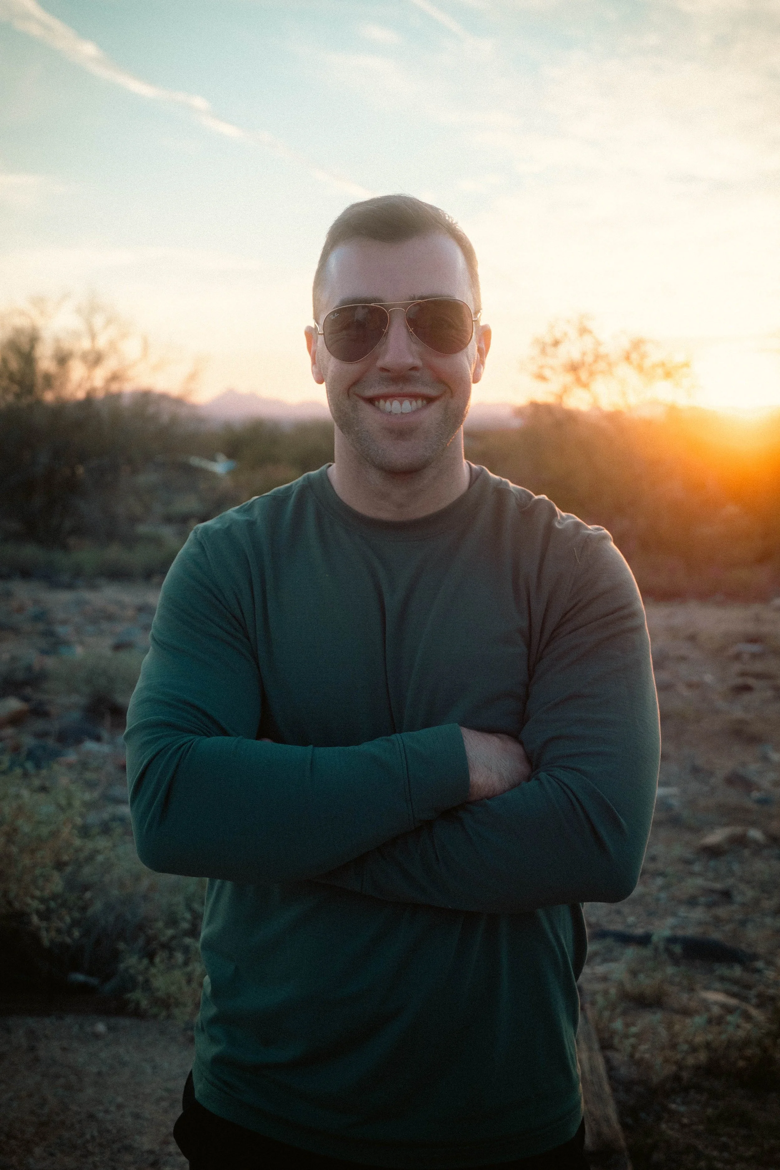 A young man with short brown hair, wearing sunglasses and a green long-sleeved shirt, smiling with crossed arms outdoors at sunset, with a desert landscape and mountains in the background.