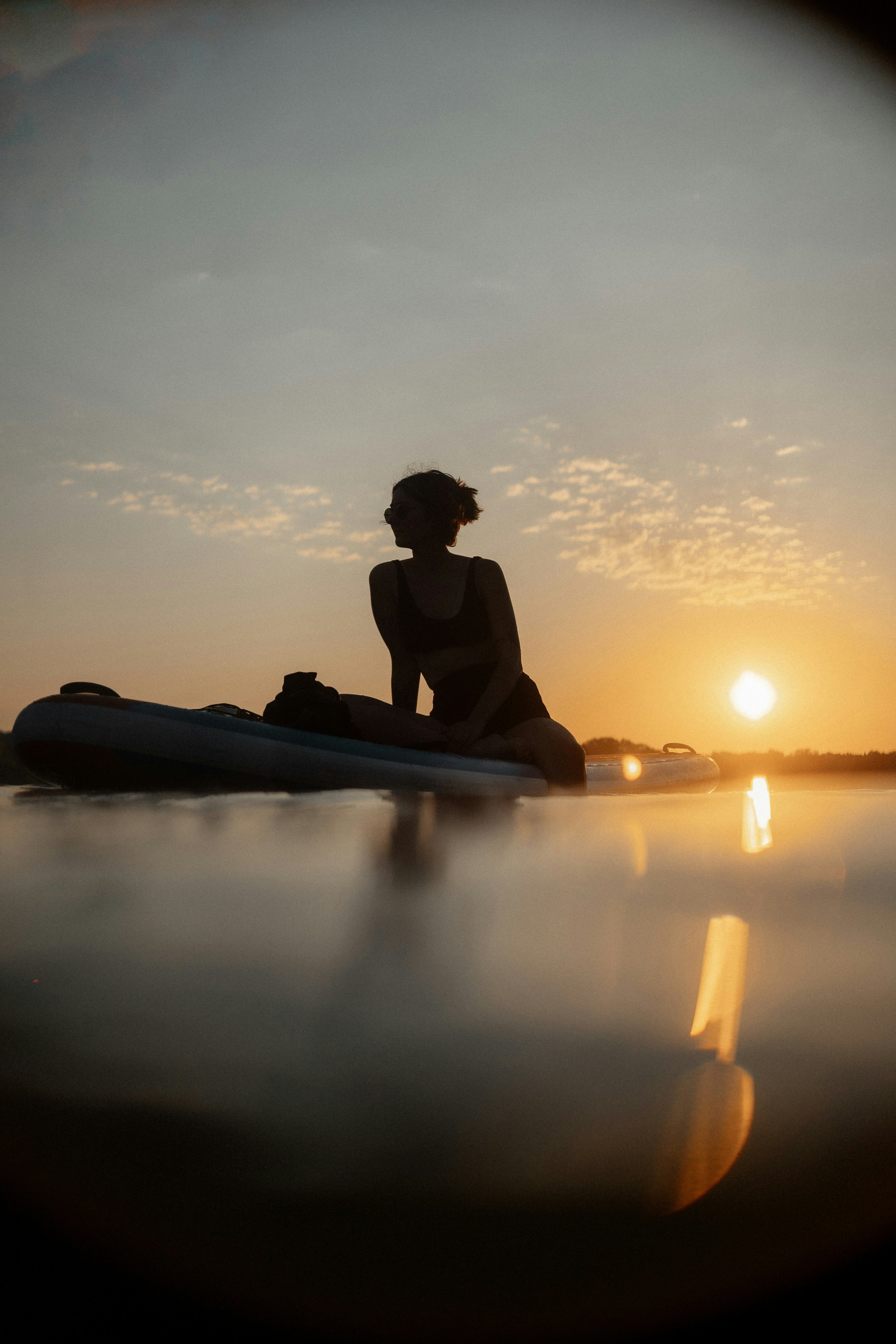 Silhouette of a person sitting on a paddleboard or kayak during sunset with a water reflection and a partly cloudy sky in the background.