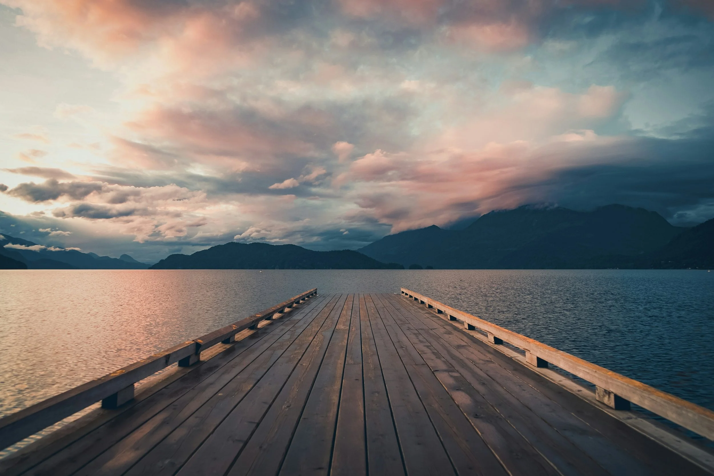 A wooden dock extending into a calm lake surrounded by mountains under a colorful cloudy sky during sunset.