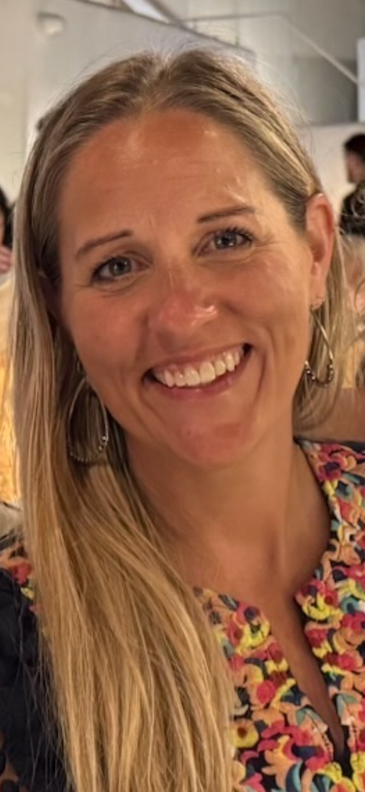 Smiling woman with long blonde hair wearing a colorful floral top and earrings in a social setting.