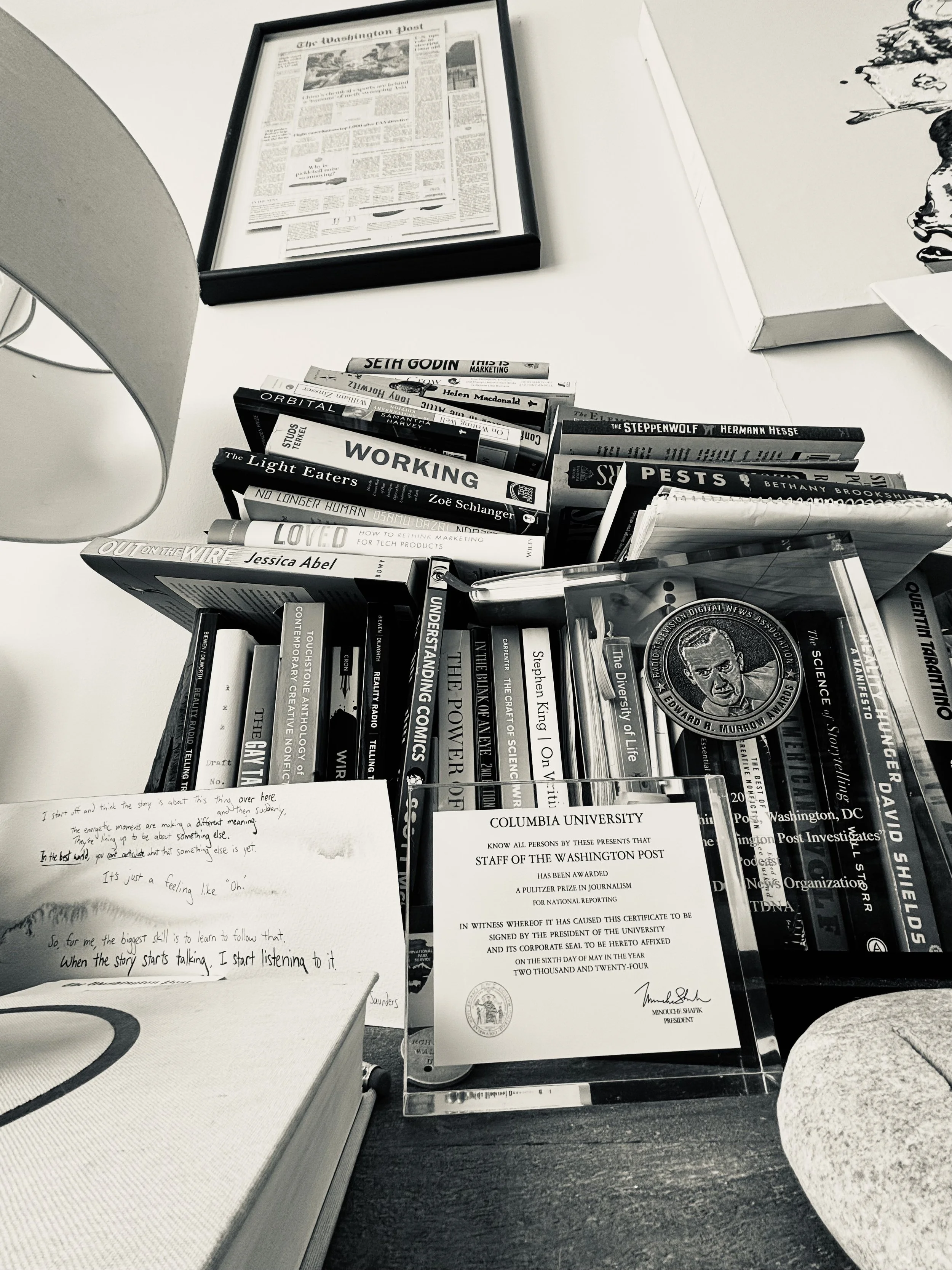 A desk with a framed newspaper, a stack of books, and certificates. The books include titles like 'Working', 'Orbital', and 'Pests'. There is handwritten note paper with text, and a small part of a chair is visible.
