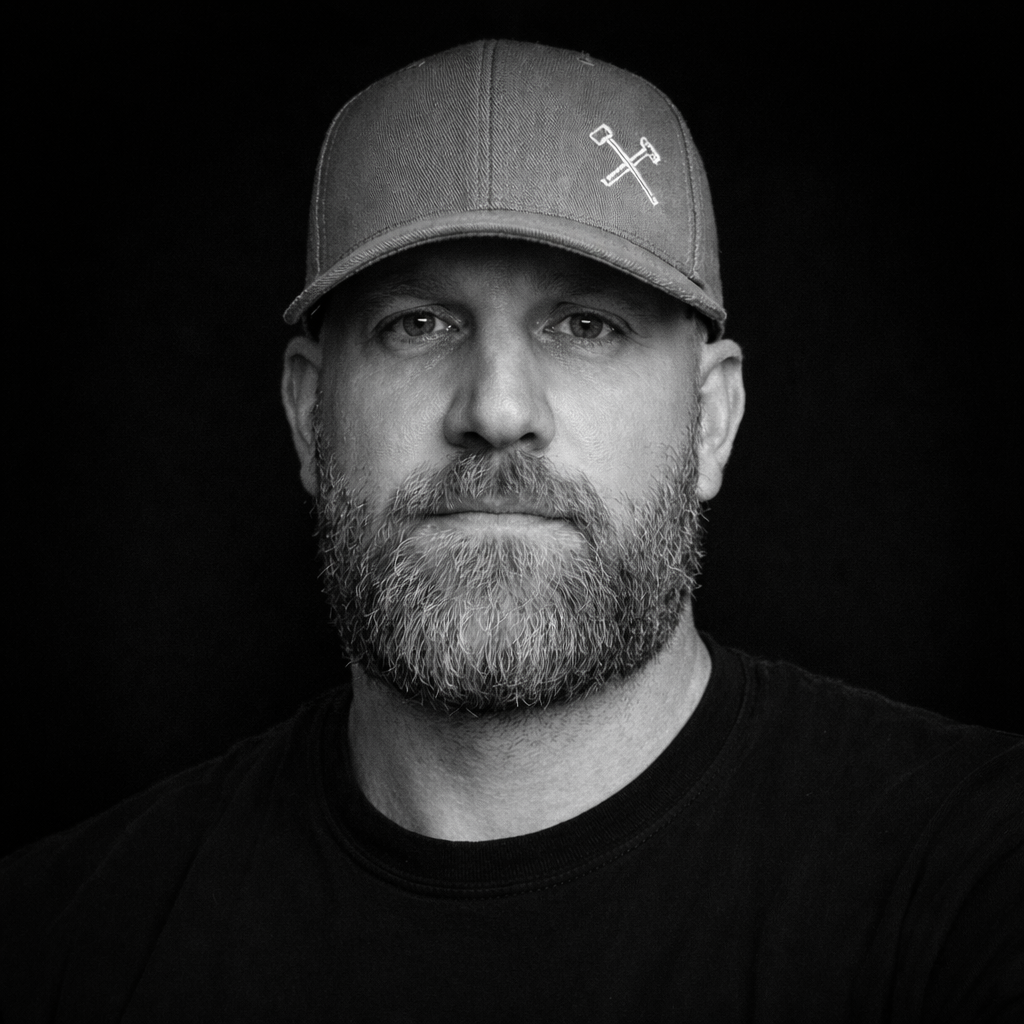 A black and white portrait of a bearded man wearing a cap with crossed tools embroidered on it, against a dark background.