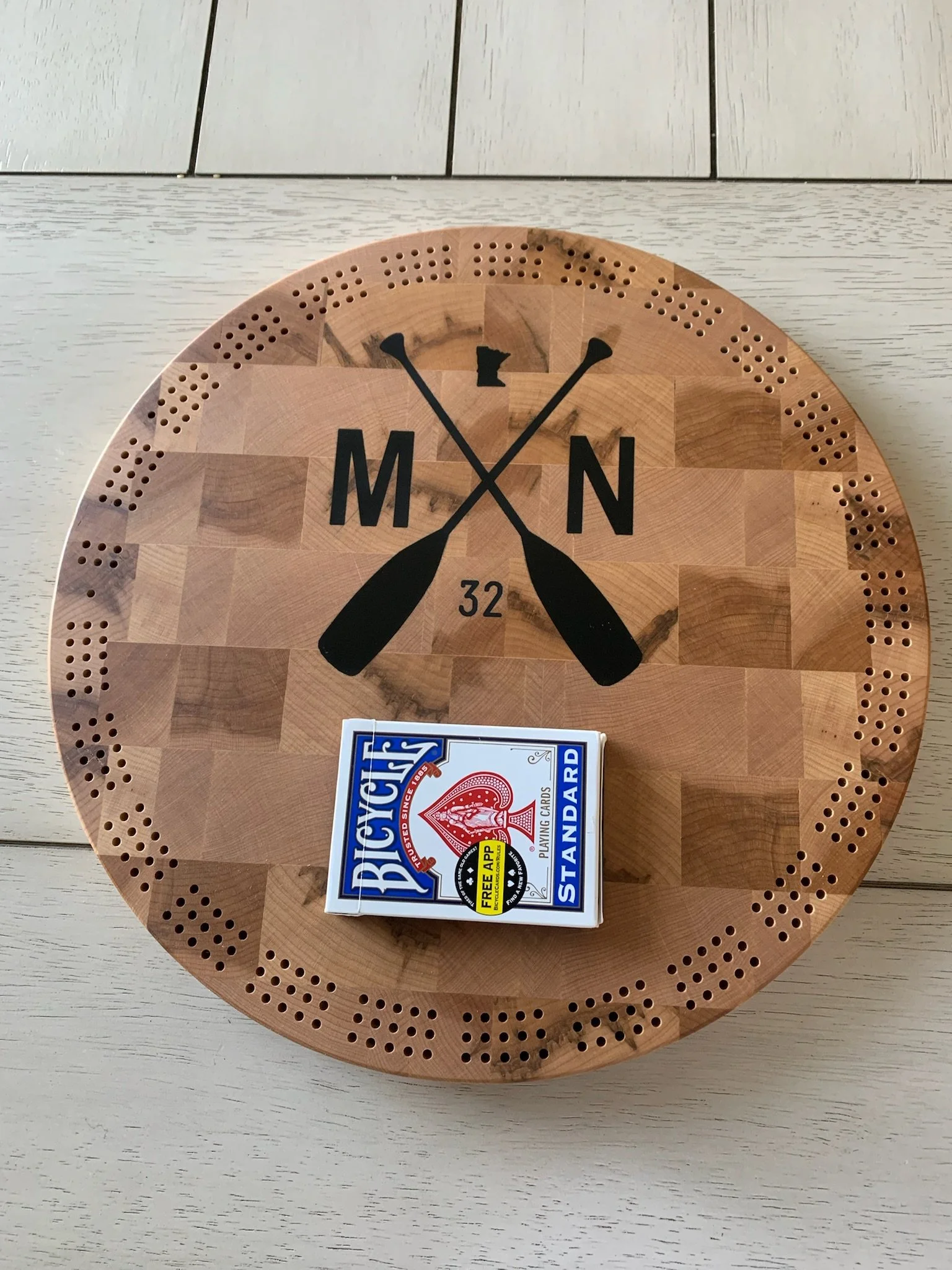 A round wooden clock with black hands and numbers, including the letters M and N, and a pack of Bicycle playing cards on a light-colored wooden table.