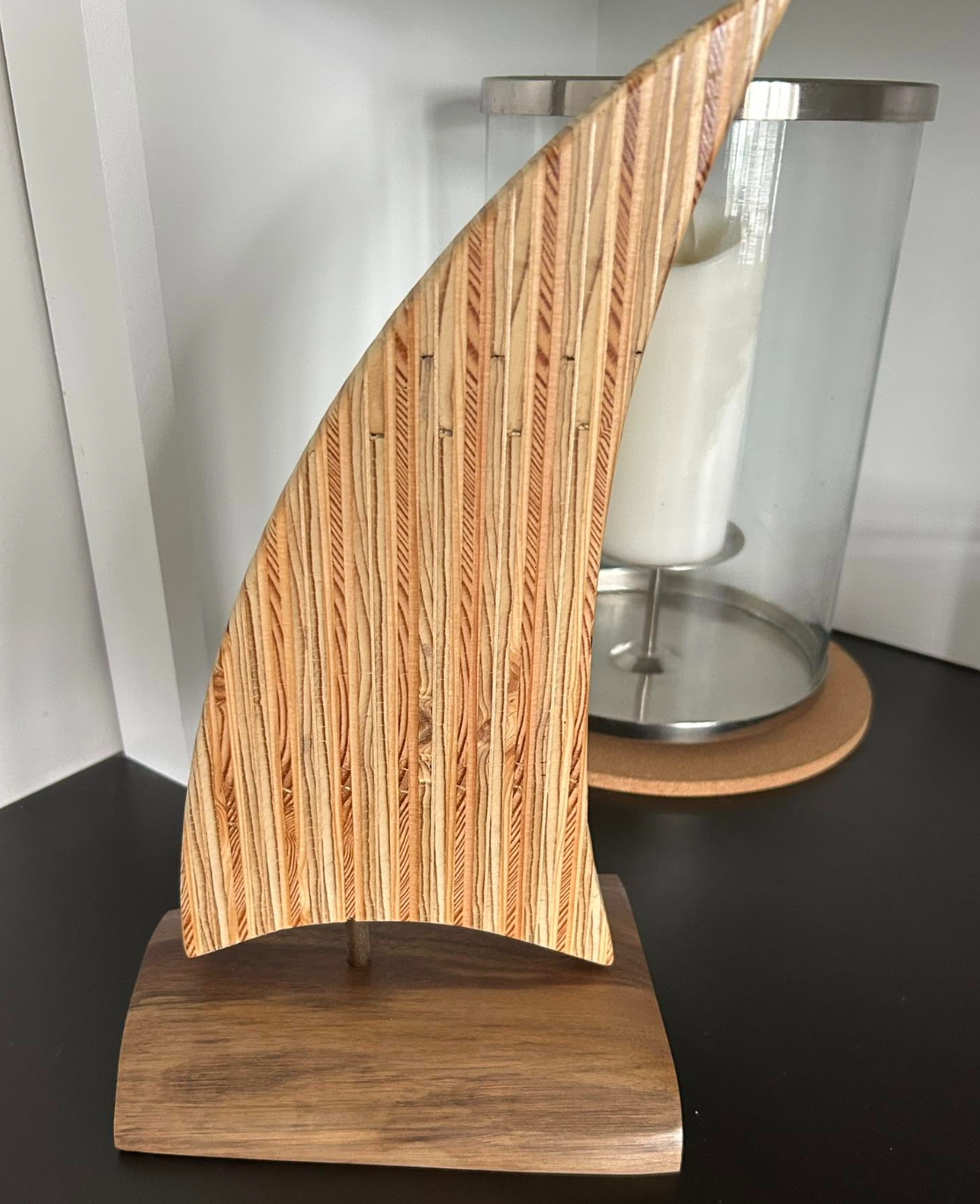 Wooden sculpture with striped grain standing on a wooden base, with a glass container filled with a white substance in the background.