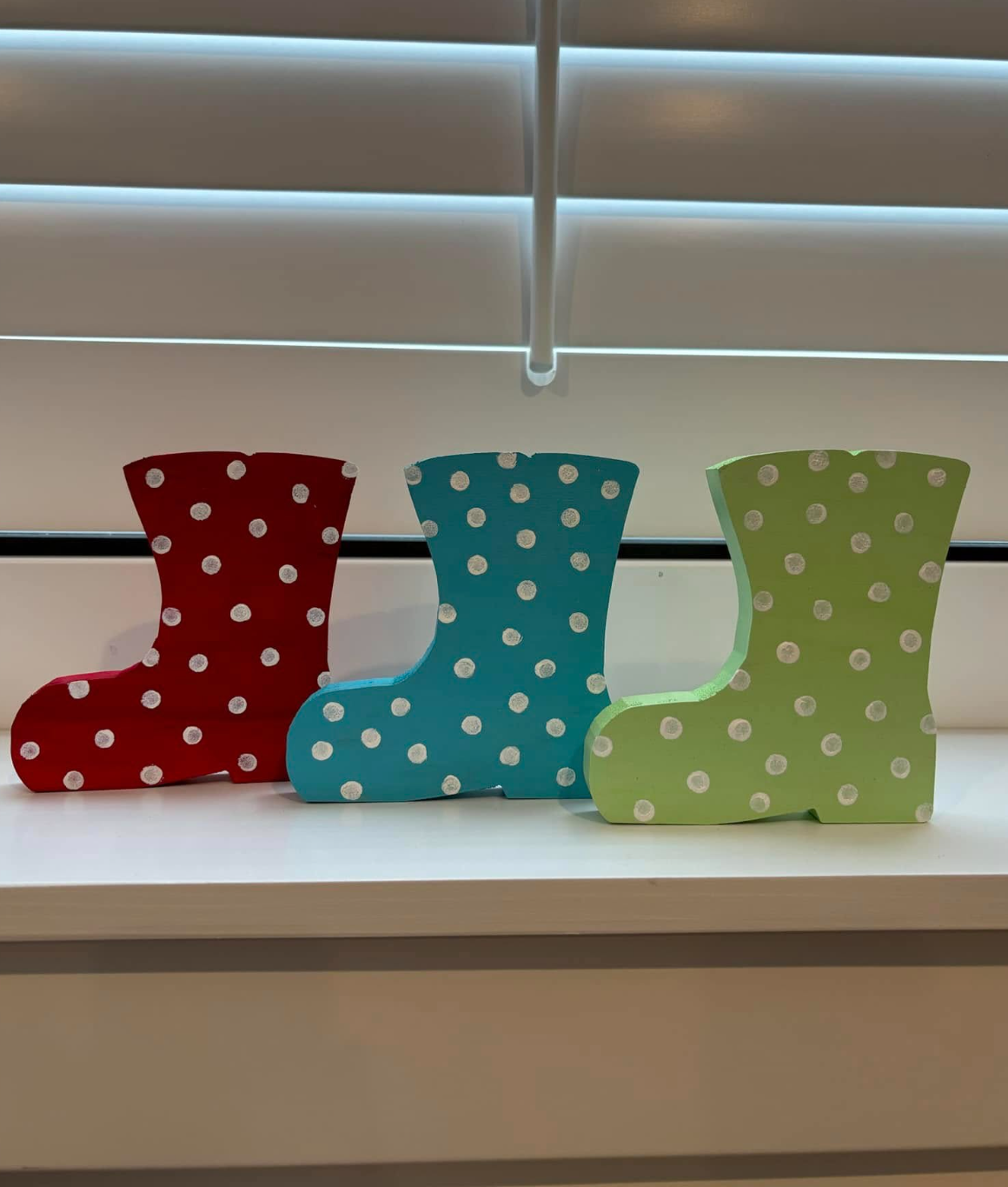 Three colorful decorative snow boots in red, blue, and green with white polka dots, lined up on a white surface under window blinds.